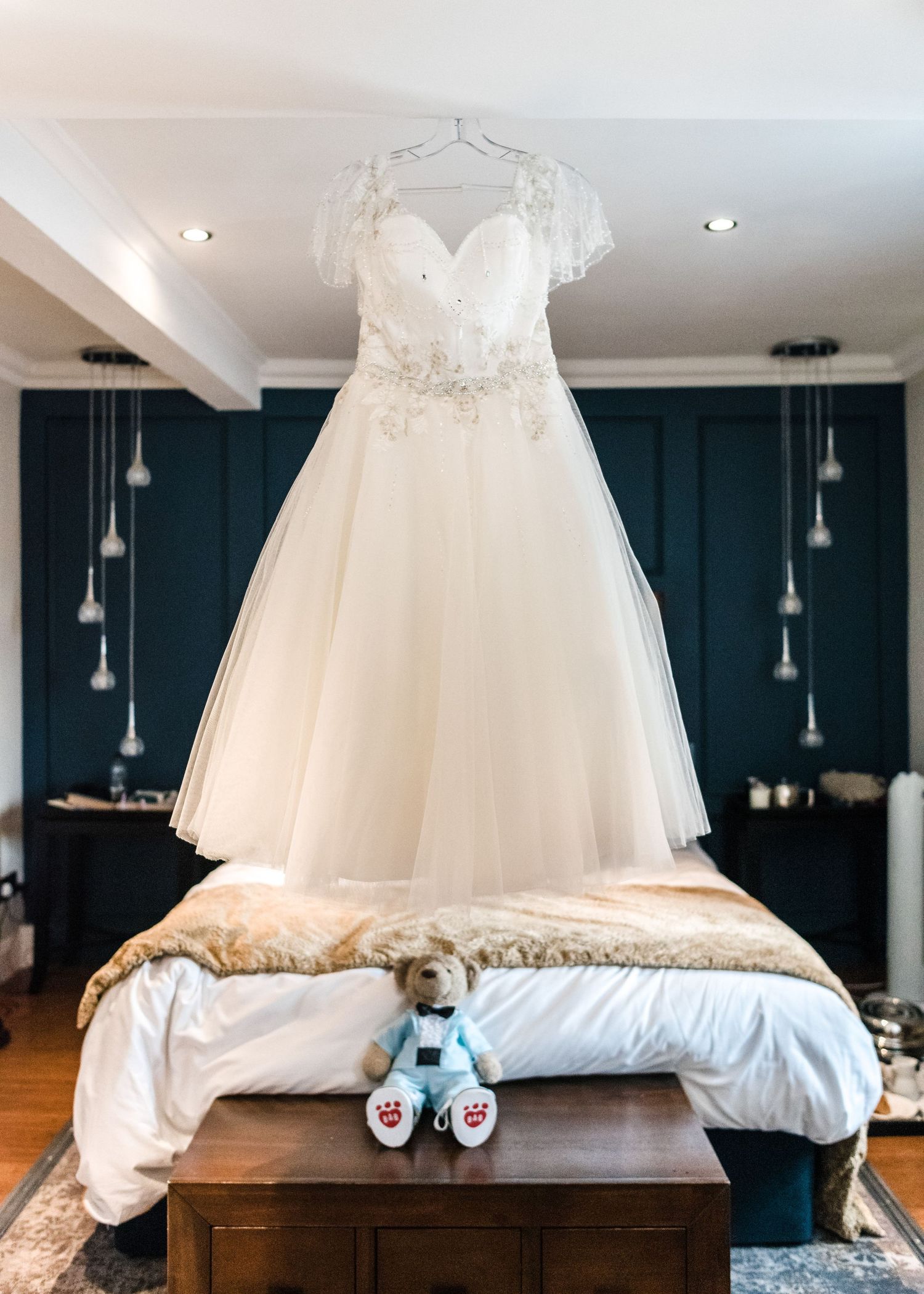 Elegant white wedding dress with lace details hanging above bed with stuffed bear.