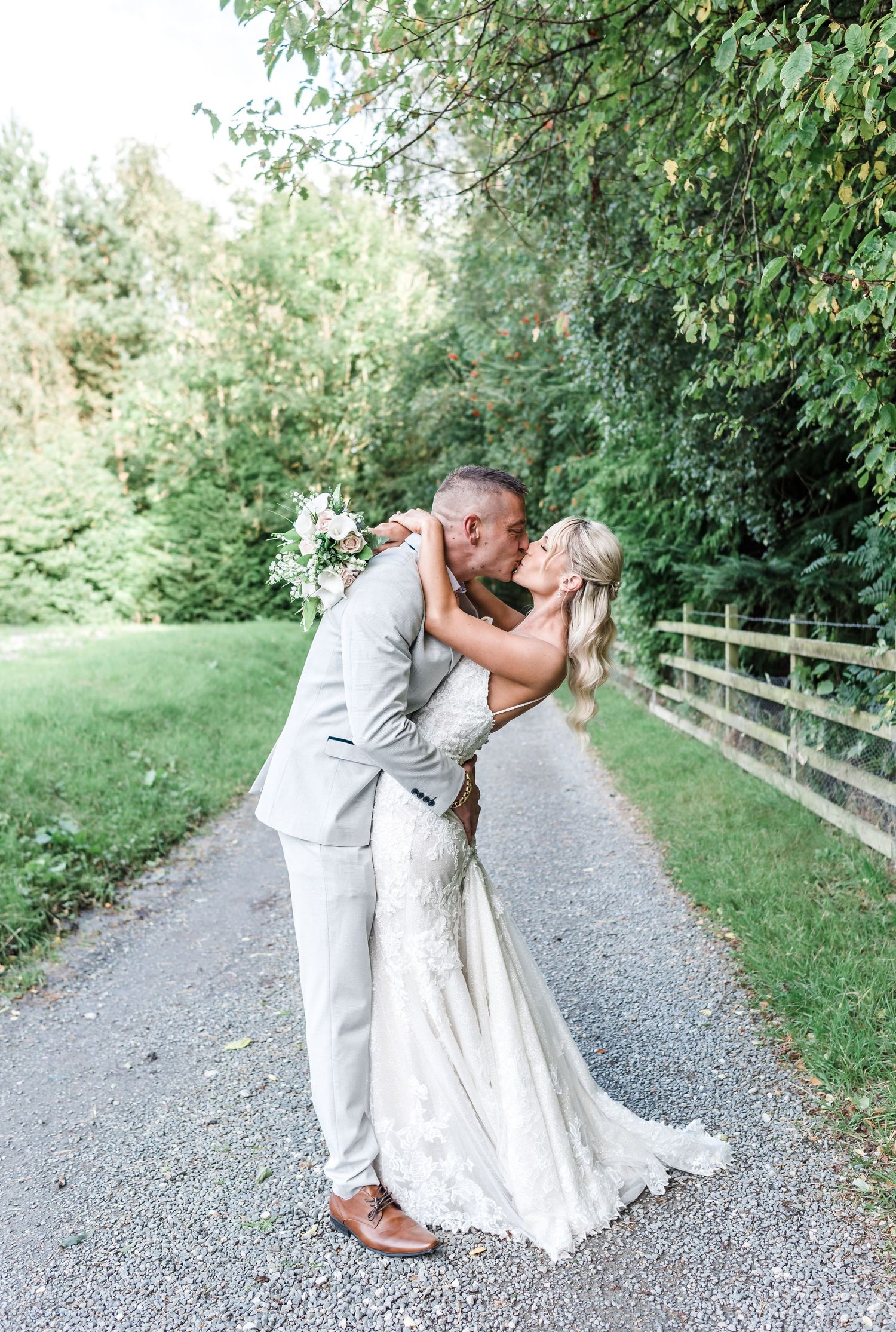 Romantic dip kiss between newlyweds on country path surrounded by greenery.