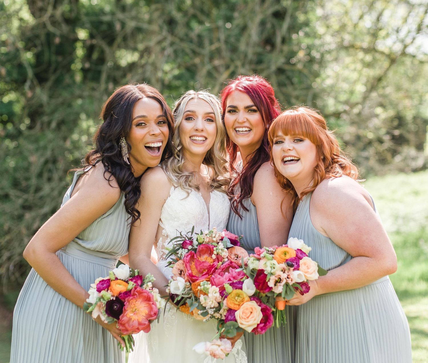 Wedding party wearing sage green bridesmaid dresses holding colorful bouquets against outdoor garden setting.