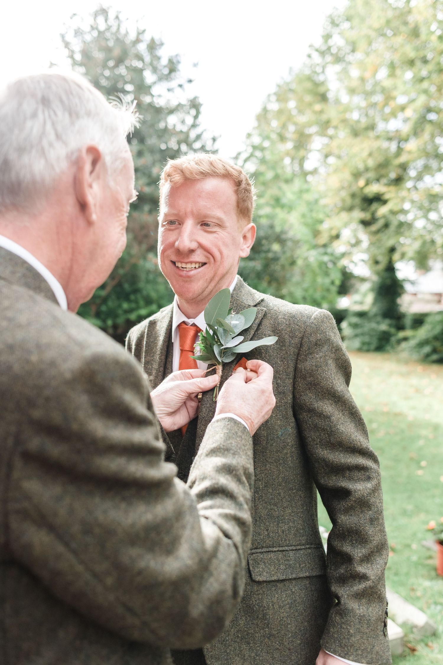A tender moment as someone adjusts a boutonniere on a tweed suit jacket outdoors.
