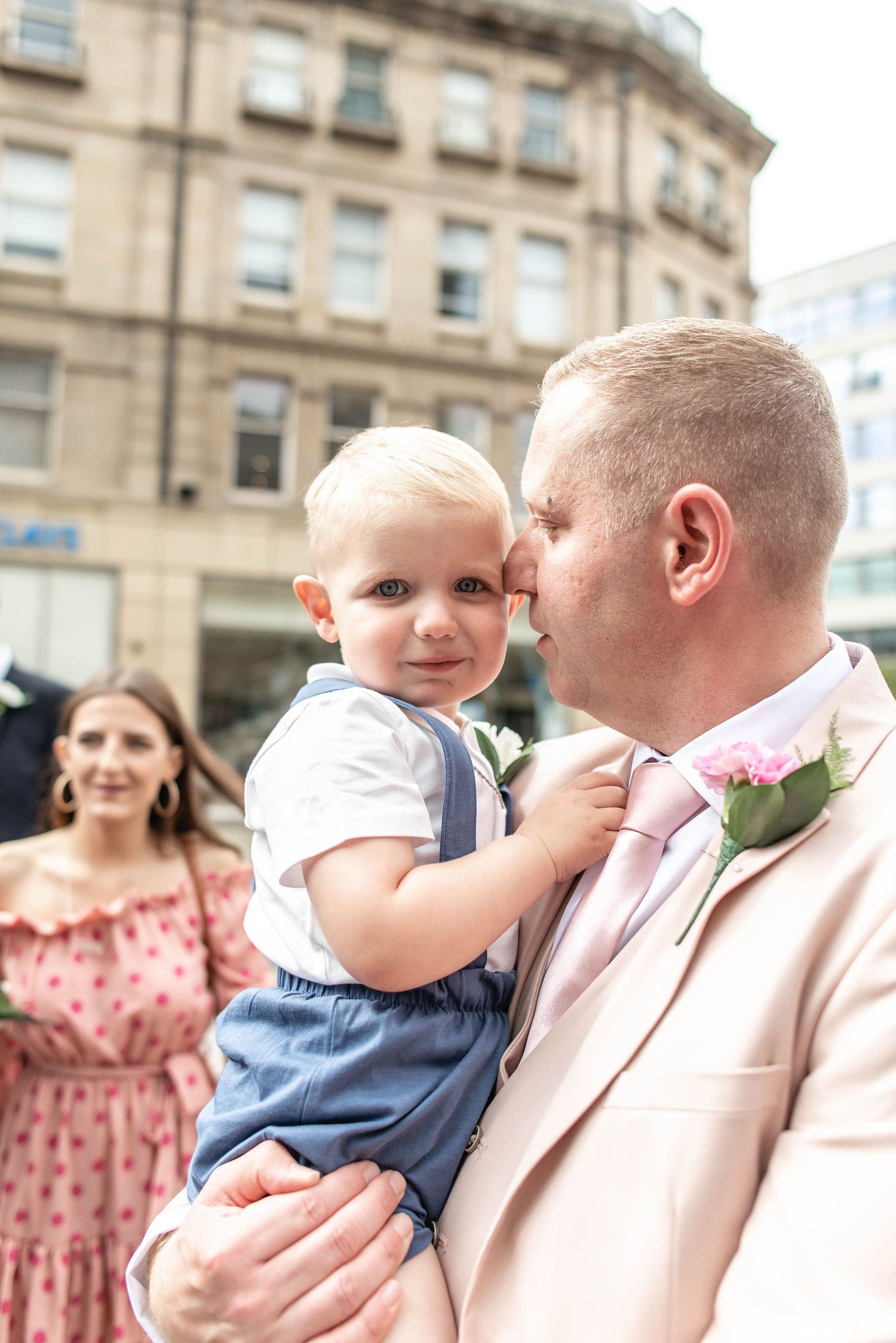 Person in pink suit holding small child in blue overalls during outdoor celebration.
