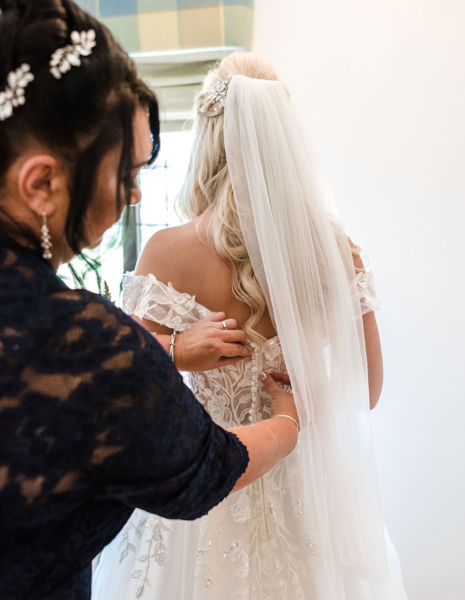 Helper adjusting buttons on back of intricate lace wedding dress with flowing veil.