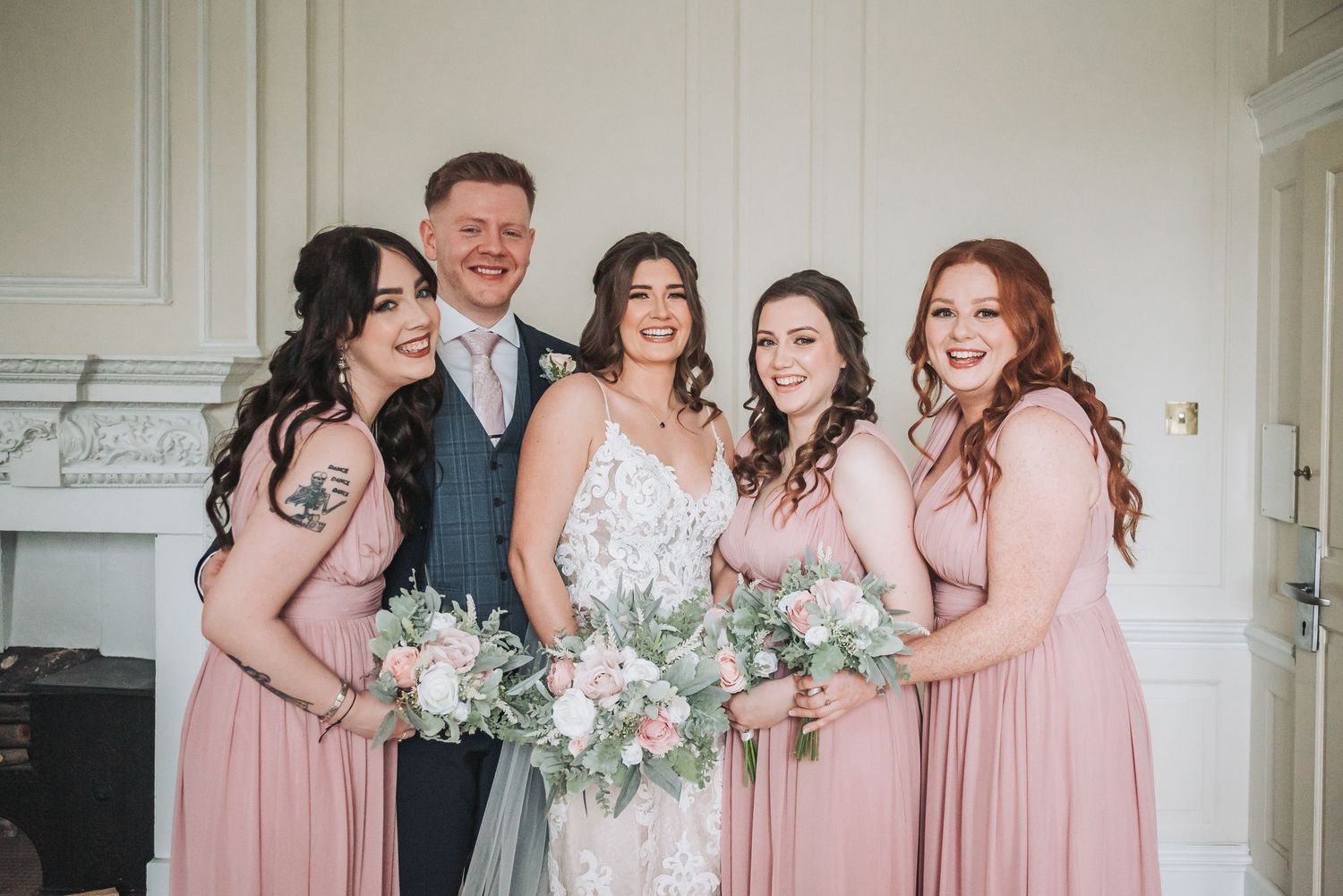 A bridal party in pink gowns and a grey vest hold white and green bouquets while smiling in front of a white interior wall.