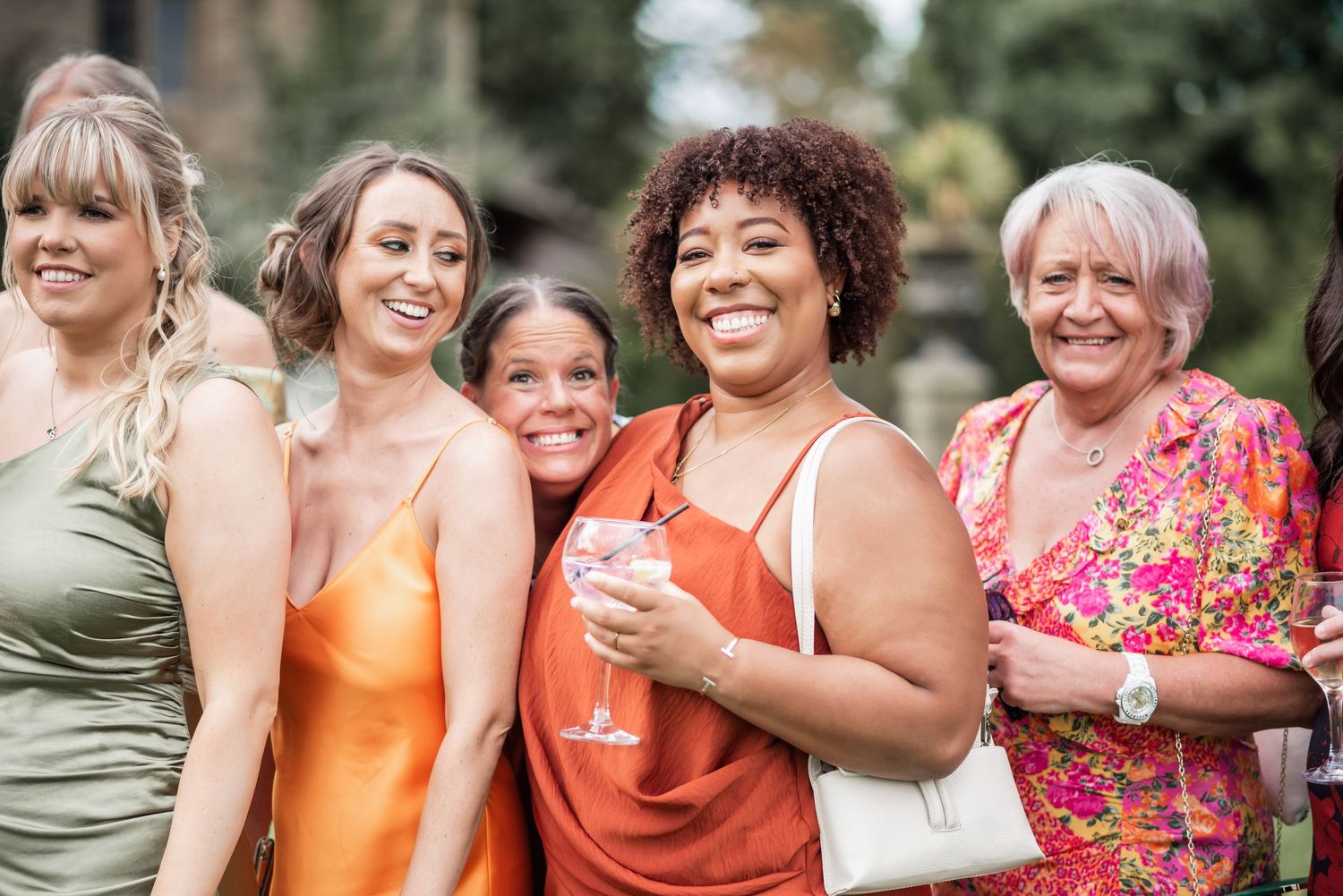 A diverse group of women in colorful dresses smile together at an outdoor gathering.