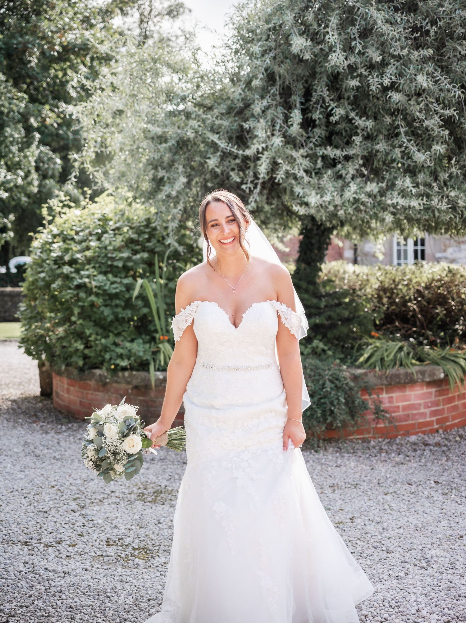A bride in an off-shoulder white wedding gown poses in a garden setting with her bouquet.