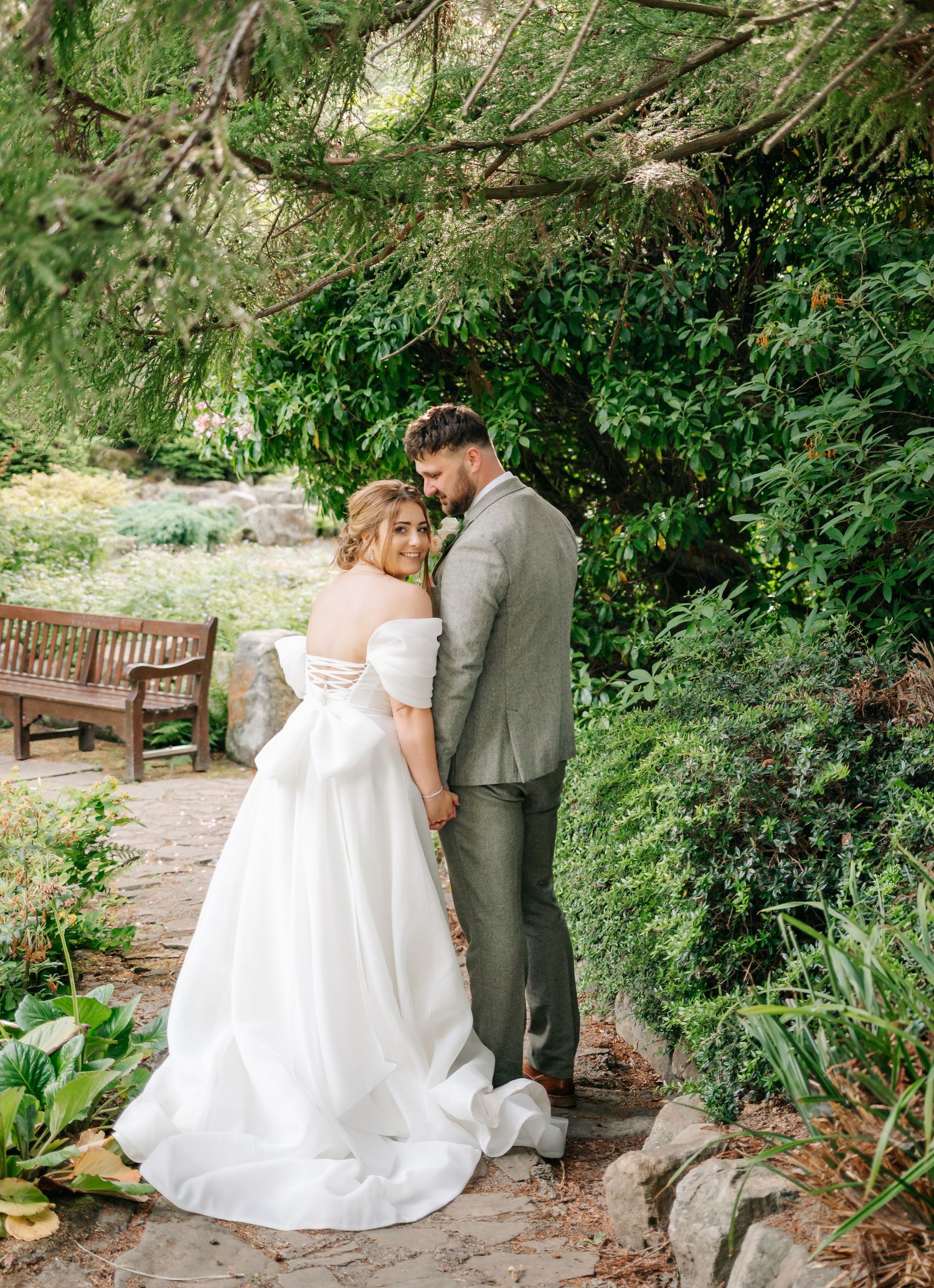 A romantic wedding couple embraces on a garden path surrounded by lush greenery.