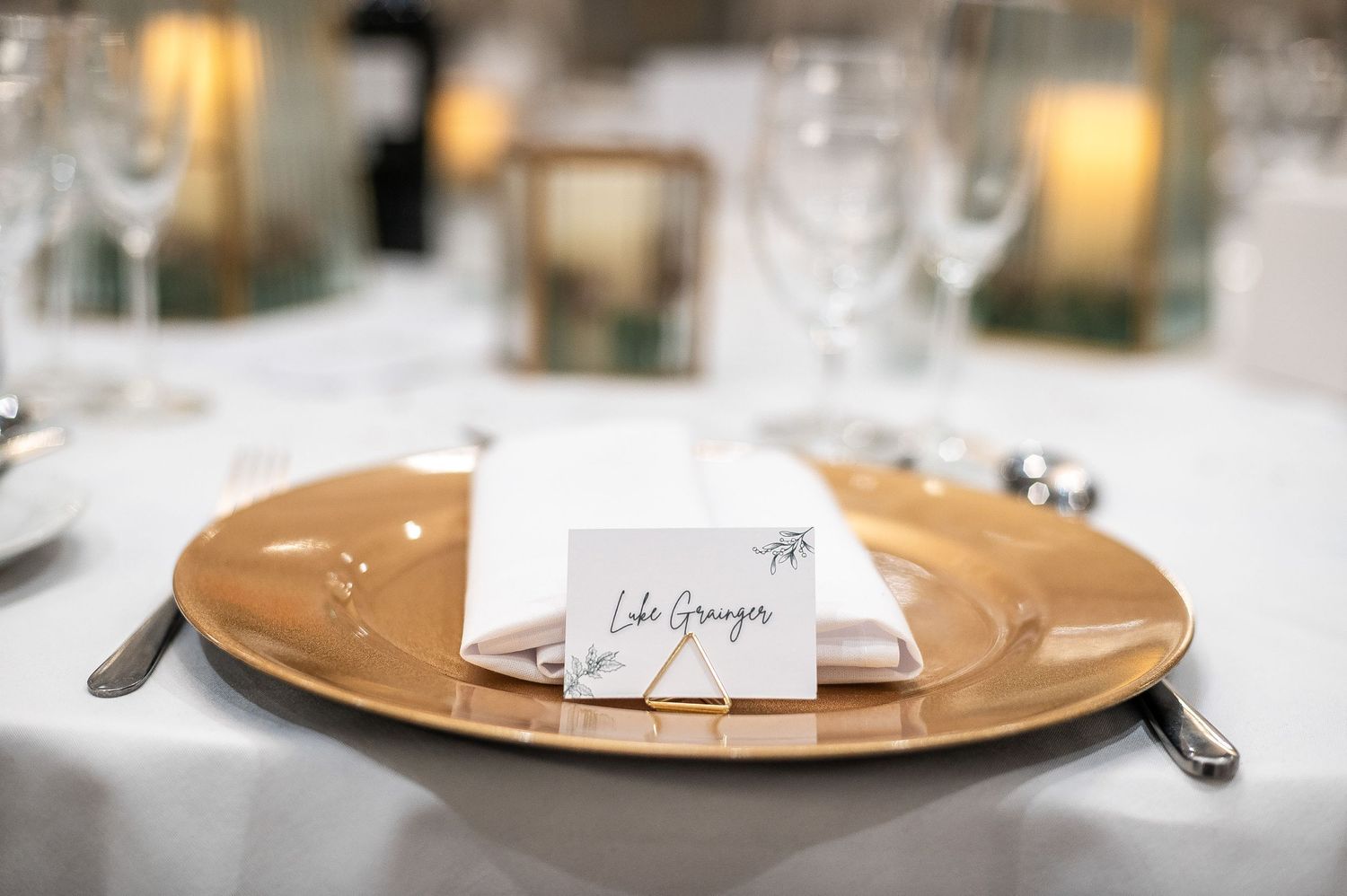 Wedding place setting with golden charger plate and name card on white tablecloth.