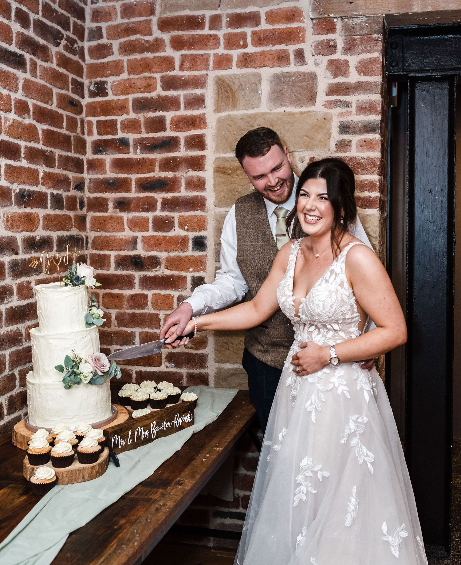 Couple cutting their elegant white wedding cake decorated with greenery at rustic brick venue.