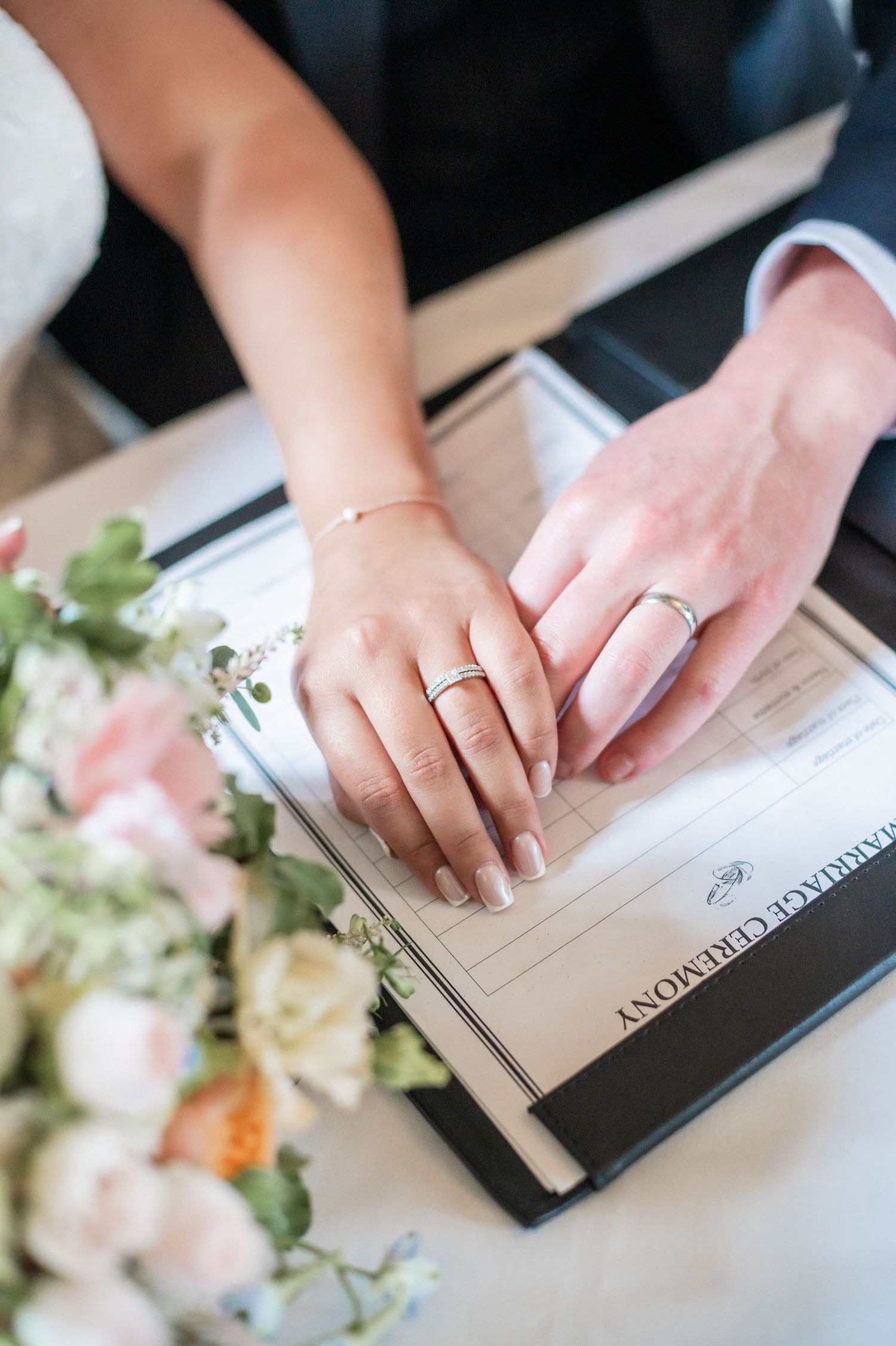 Close-up of hands with wedding rings signing marriage documents next to delicate floral arrangements.