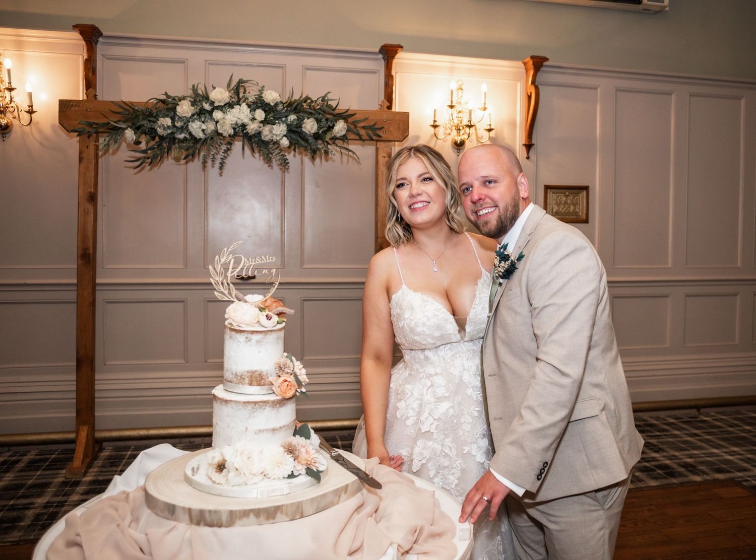 A couple poses together by their three-tiered white wedding cake during their reception.
