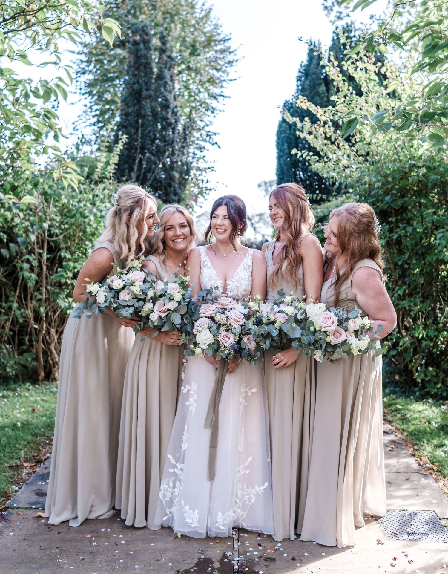 Bridesmaids in flowing champagne gowns hold lush white bouquets while laughing together in a garden setting.