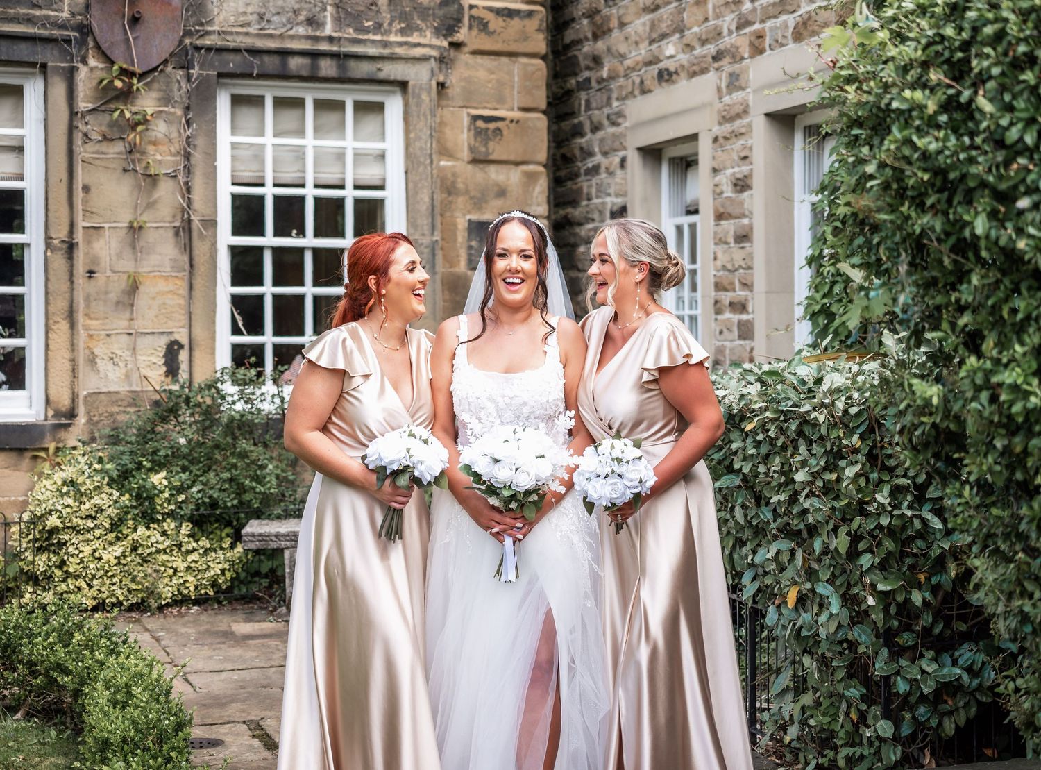 Three bridesmaids in champagne dresses posing with bouquets outside stone building.