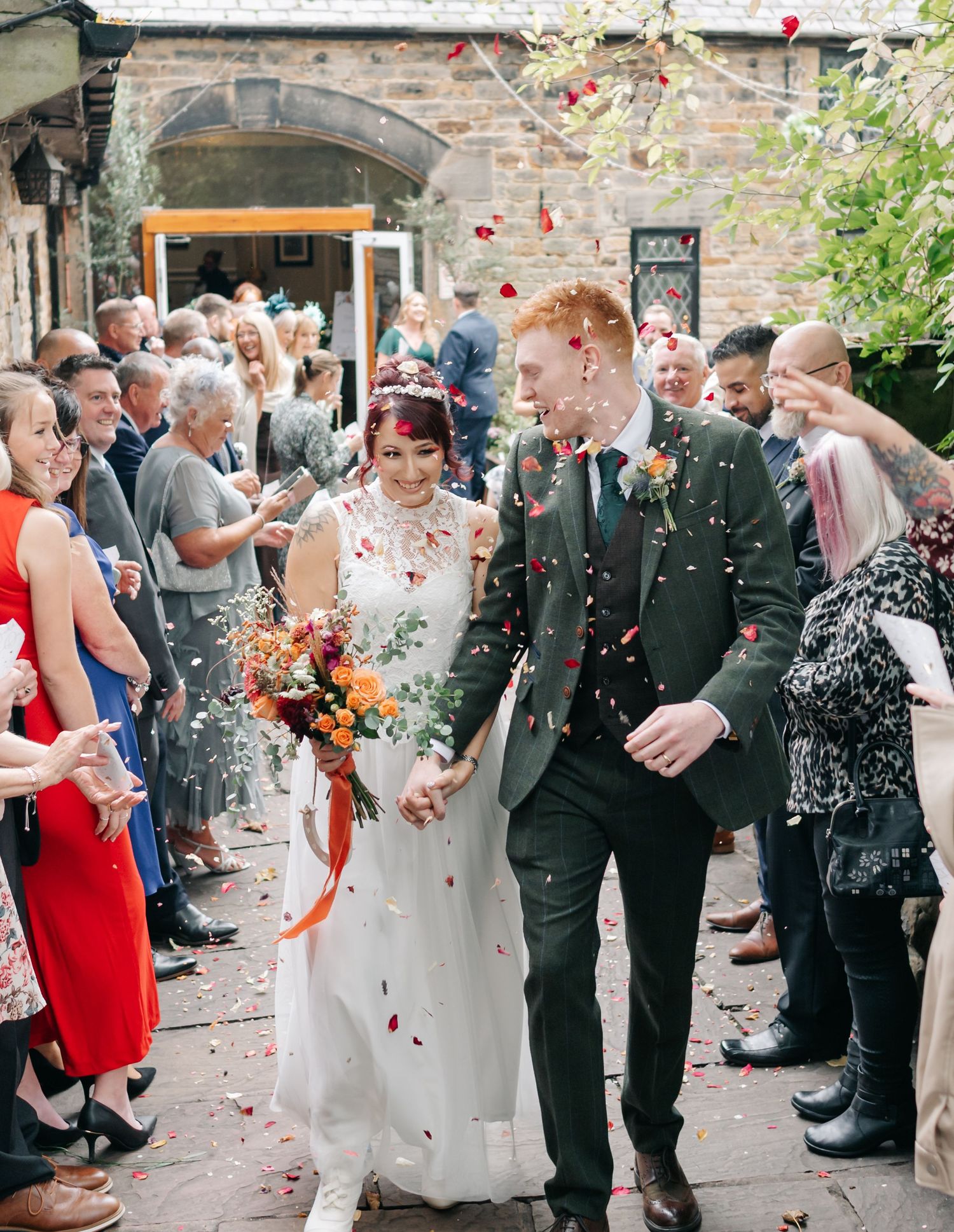 Wedding party walks through shower of confetti outside brick venue with guests lining the path.