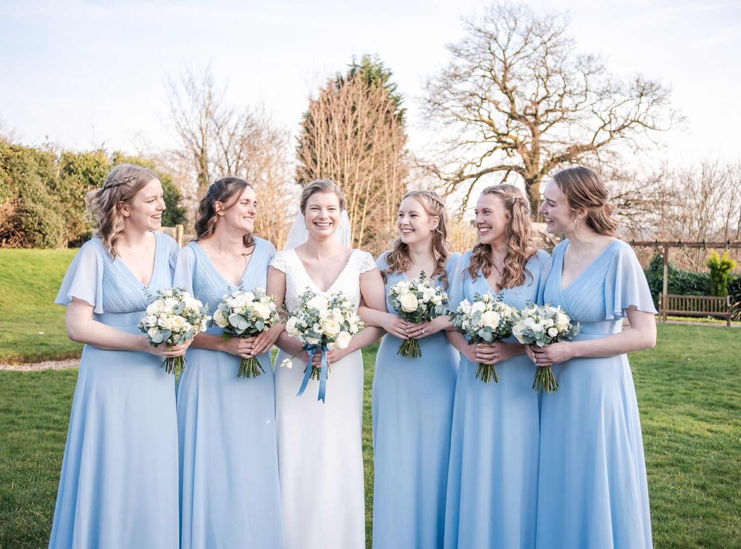 Bridesmaids in light blue dresses stand together holding white floral bouquets during an outdoor wedding ceremony.