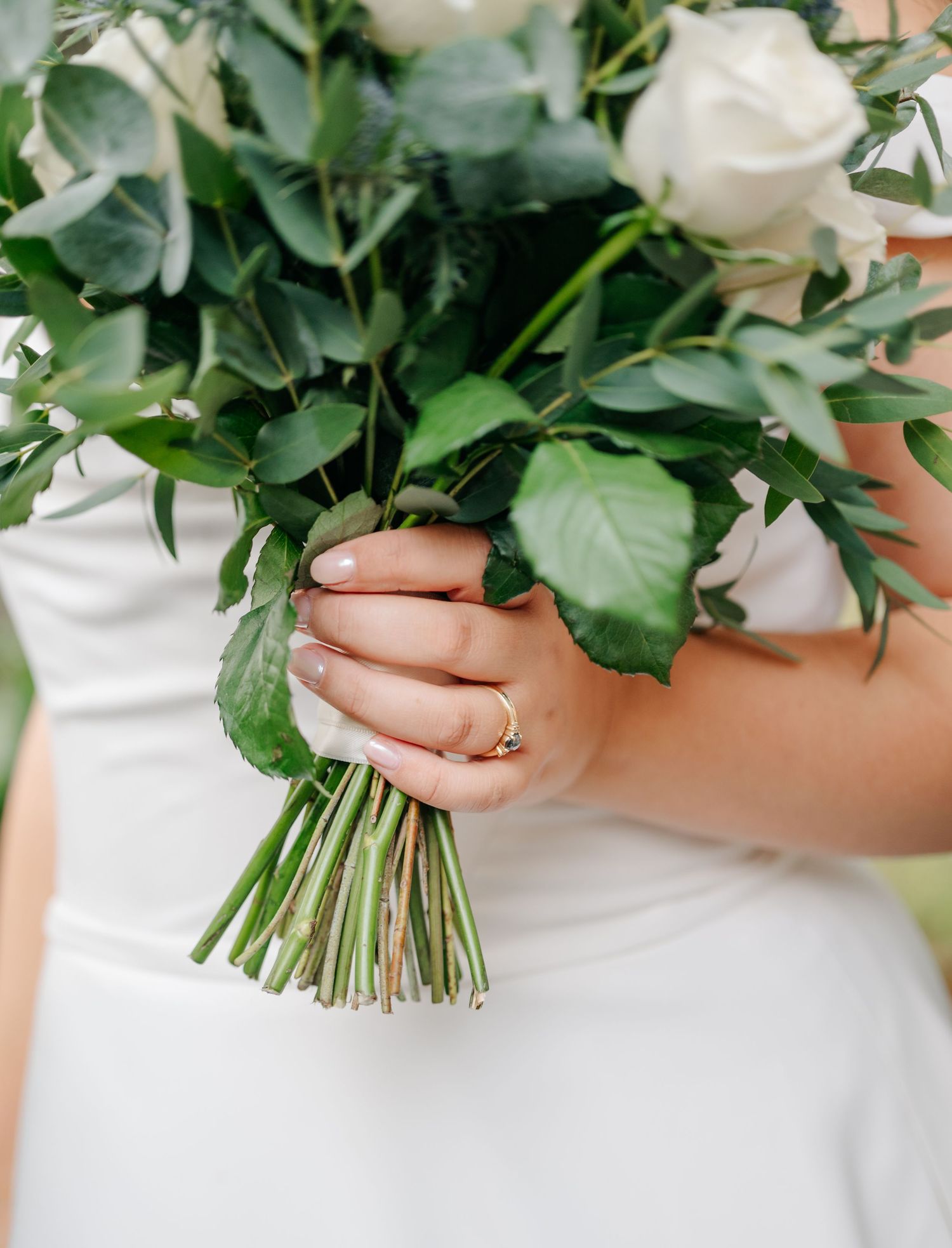White rose wedding bouquet with green foliage held against a white dress.