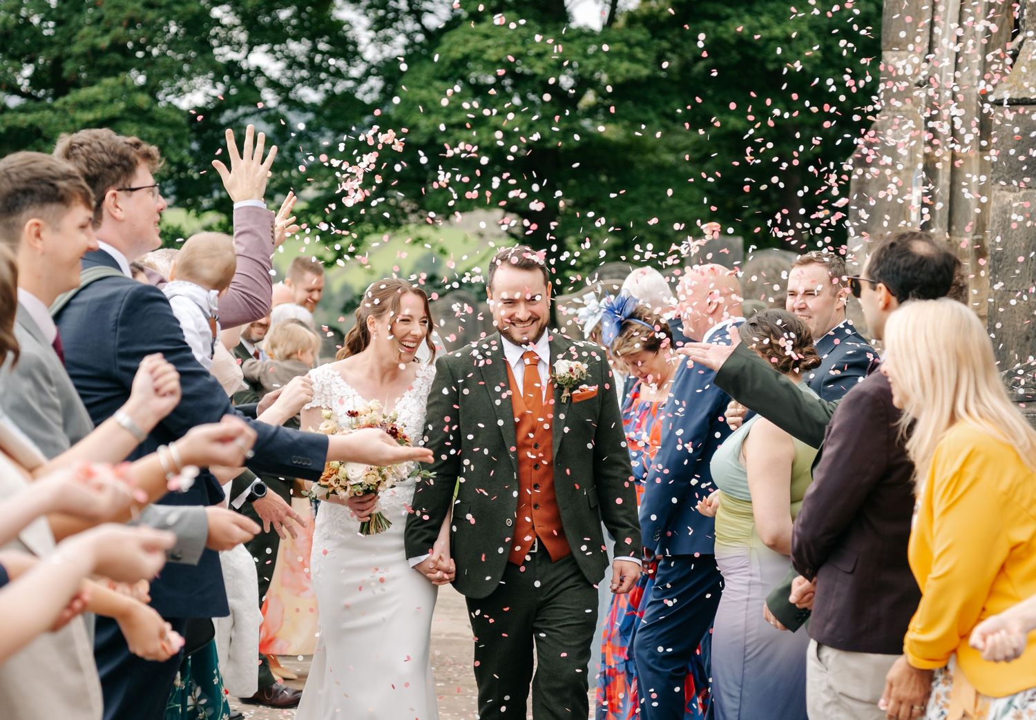 Smiling newlyweds hold hands and walk through a crowd of cheering guests throwing confetti outside a wedding venue.
