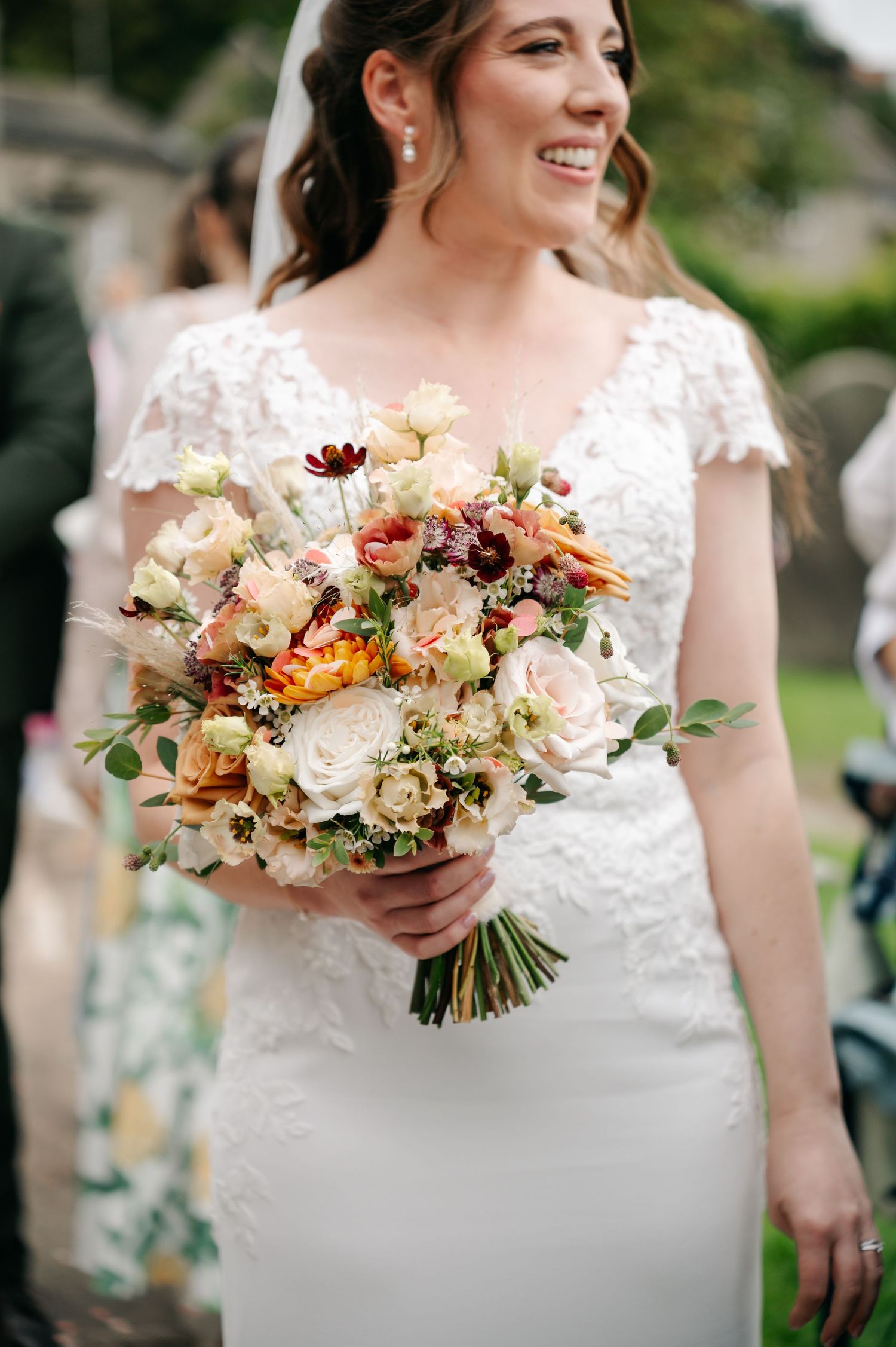 Smiling bride in a lace cap-sleeve wedding dress holds a romantic bouquet of cream, peach and orange roses.