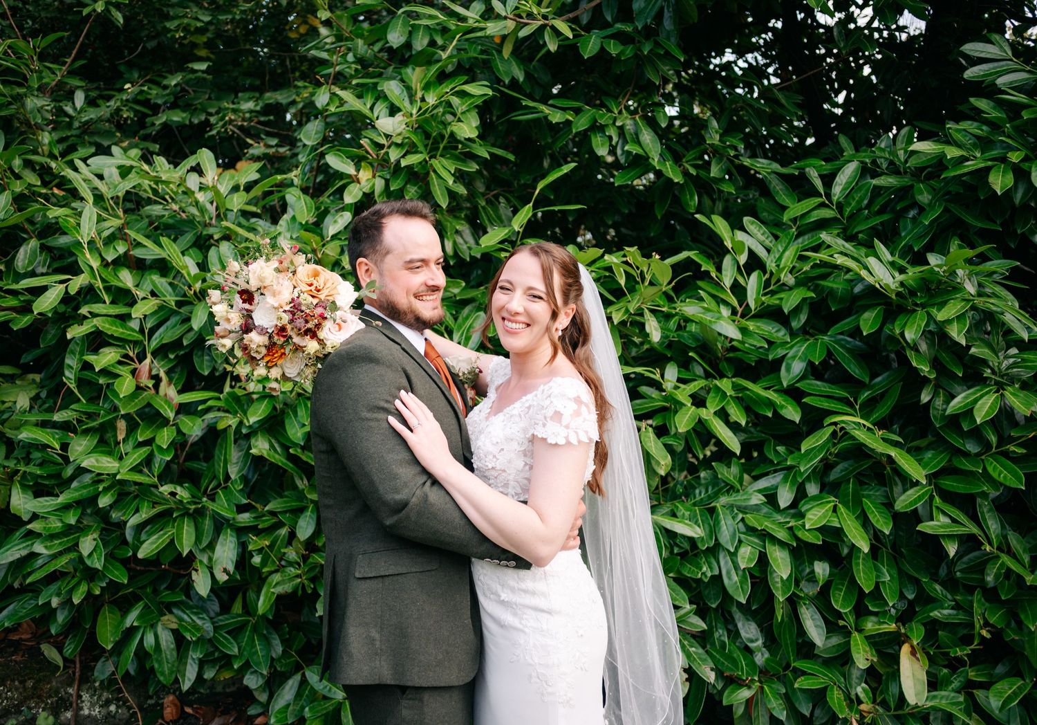 Smiling bride in a lace wedding dress and veil embraces her groom in an olive suit, holding an autumn bouquet before lush green foliage.