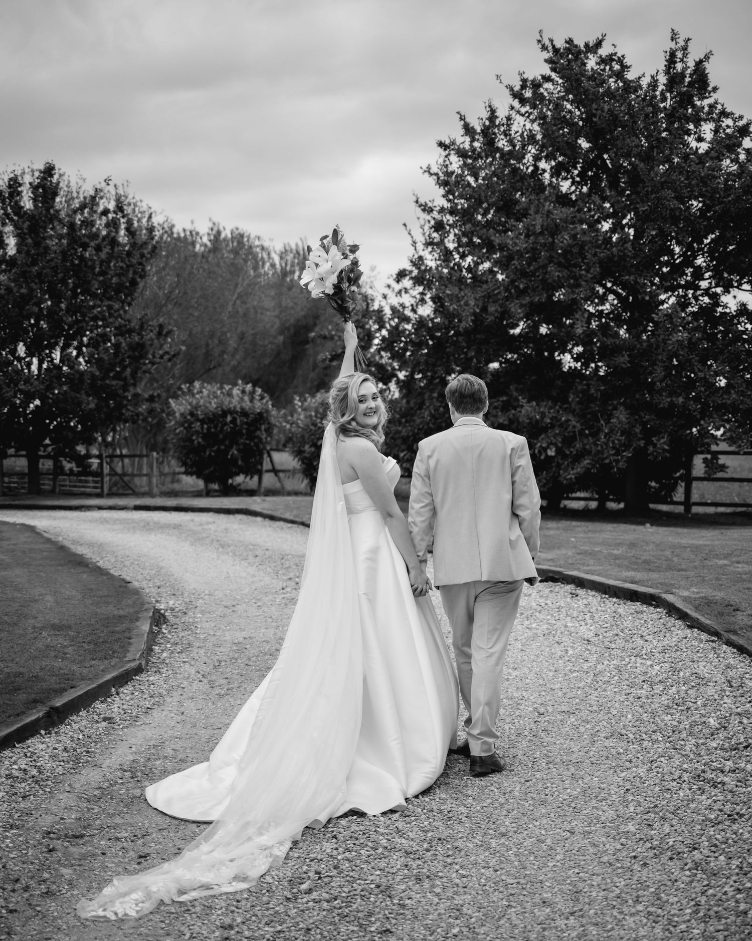 A couple in wedding attire walks along a gravel path surrounded by trees in black and white photography.