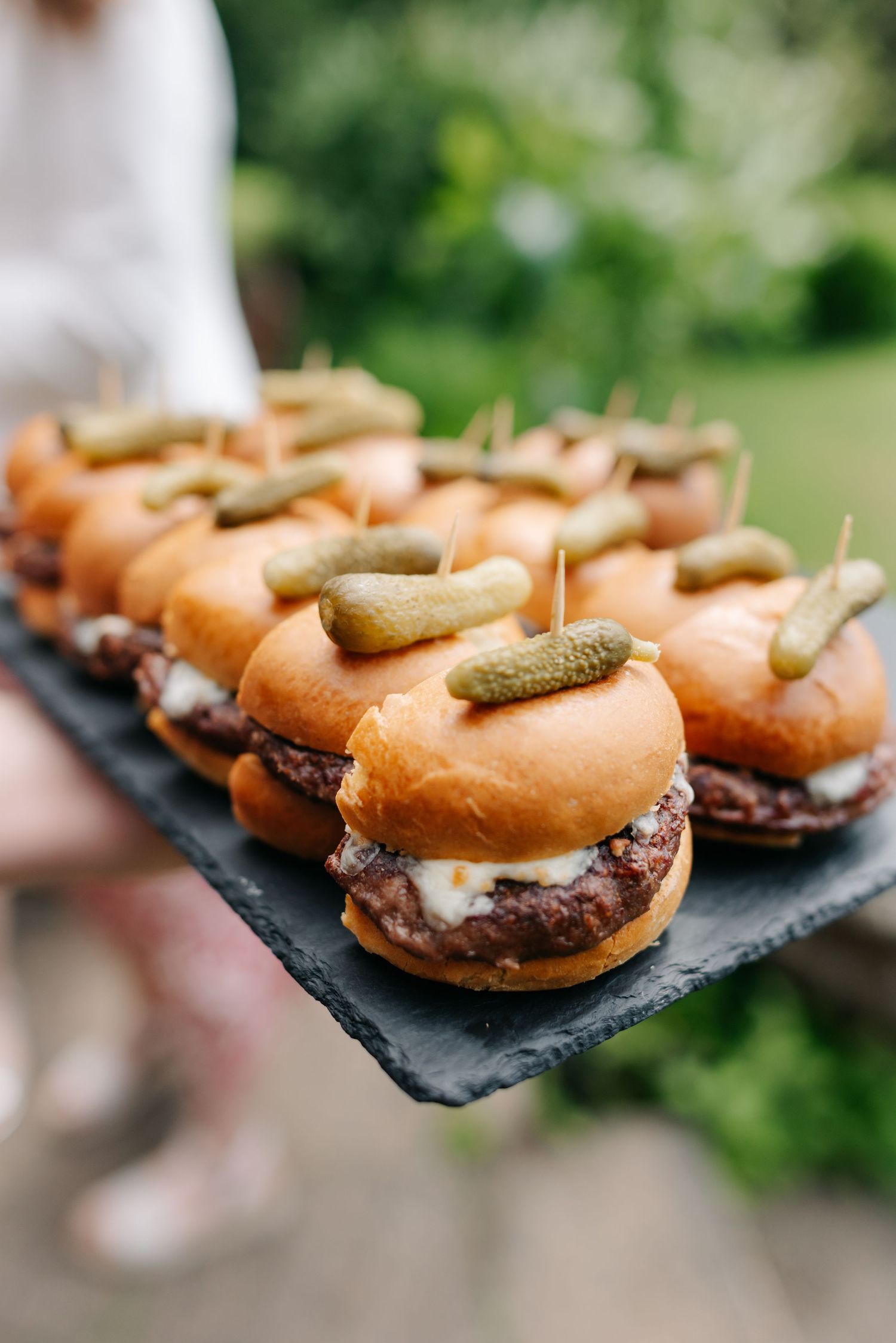 Mini burgers with pickles served on black slate platters at a wedding reception.