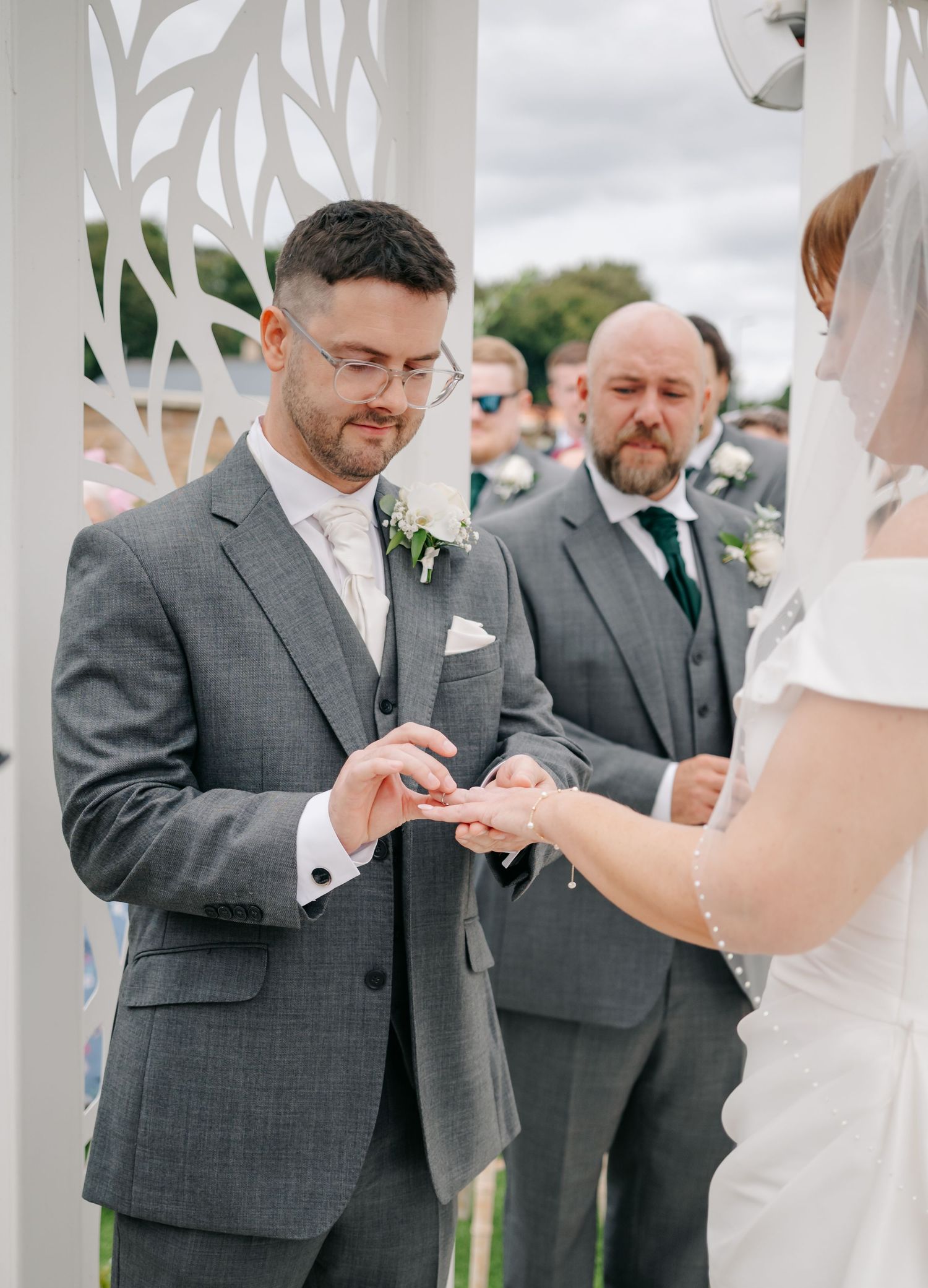 Wedding ceremony participants exchanging rings under a white decorated pergola.