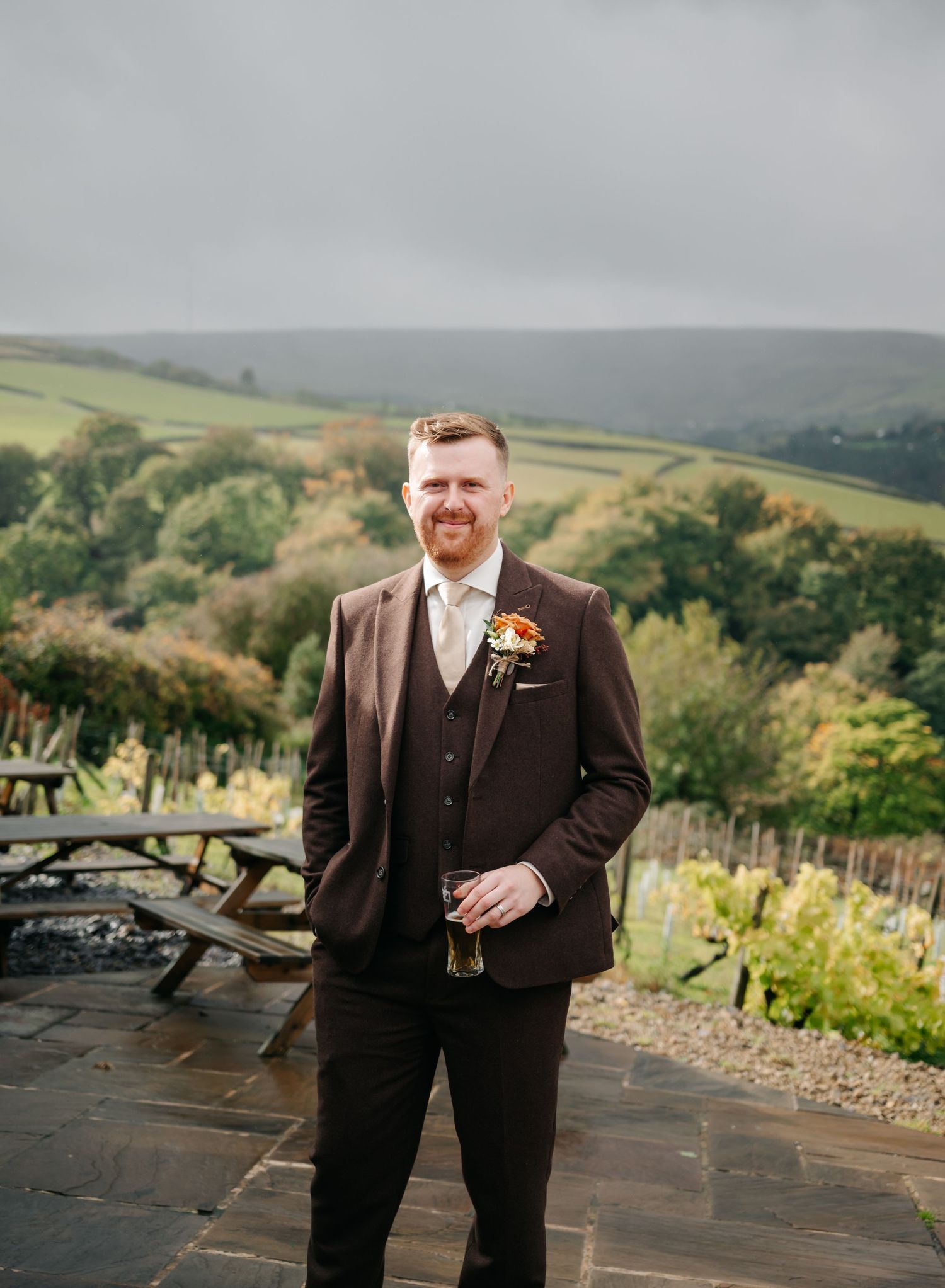 Groom in a brown suit poses outdoors against a scenic countryside backdrop with rolling hills.