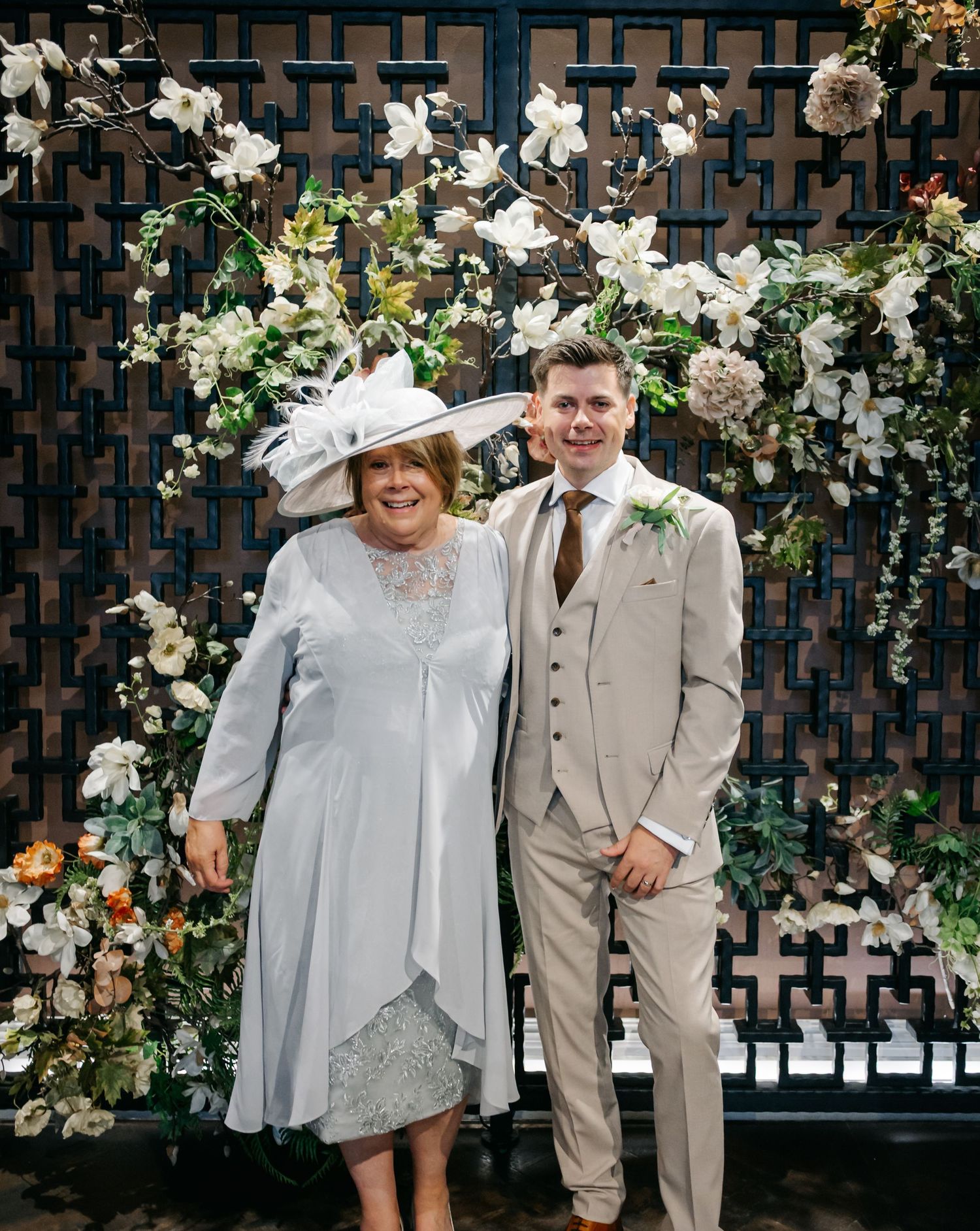 Wedding party poses against dark lattice wall decorated with white floral arrangements.