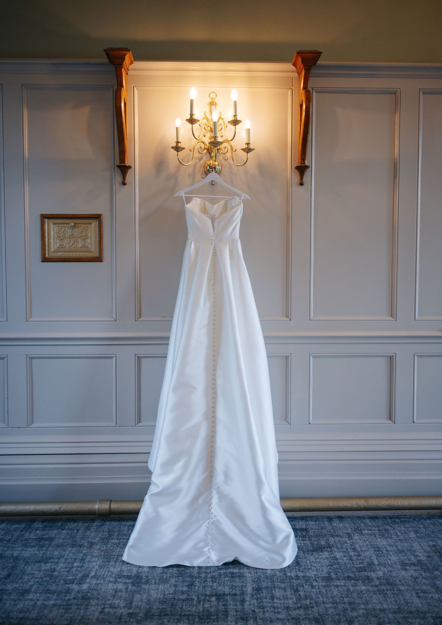 Elegant white wedding dress hanging on display against a gray paneled wall with sconce lighting.