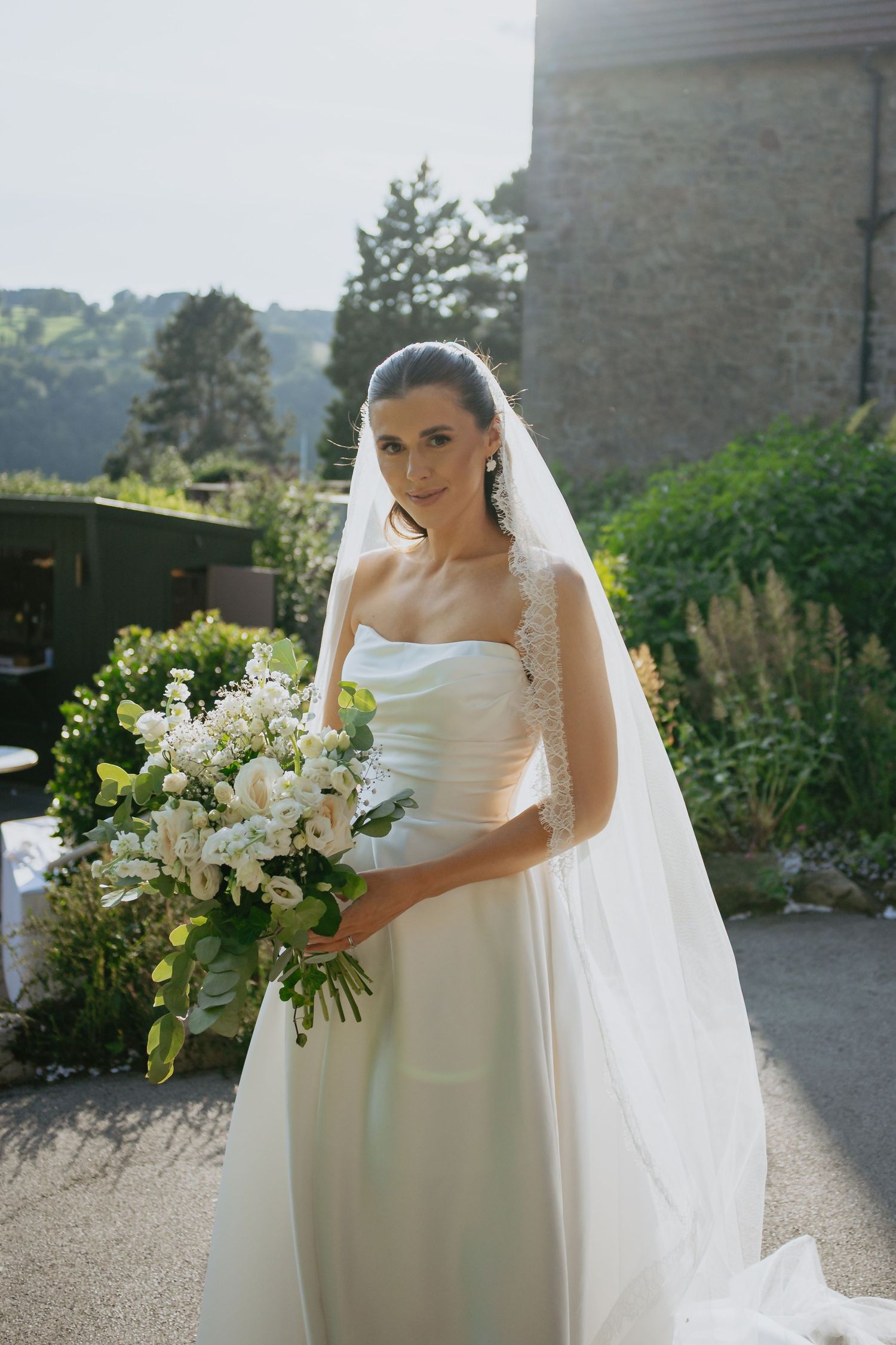 A bride in an elegant white gown holds a lush bouquet of white flowers against a stone building backdrop.