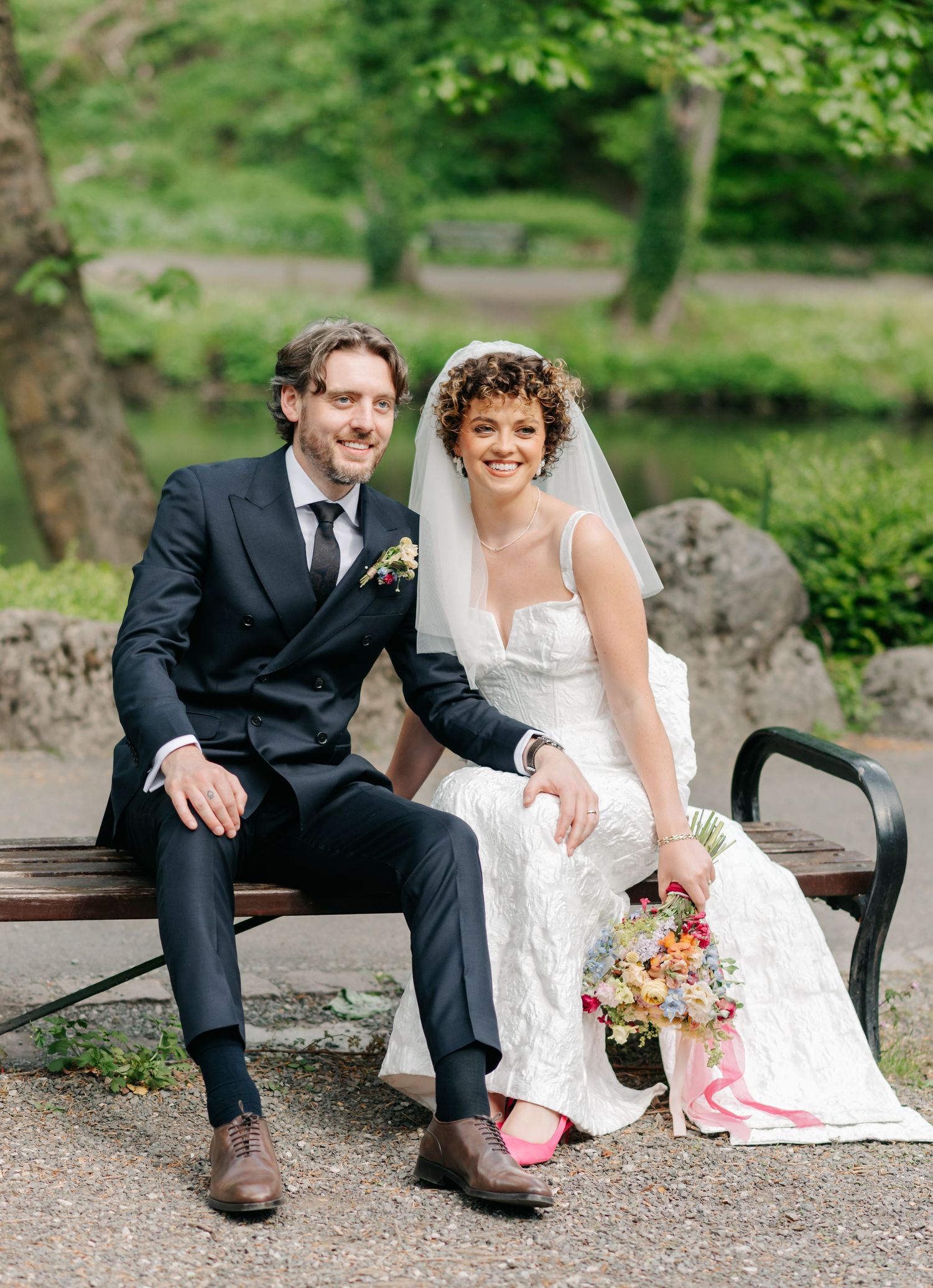 Wedding couple sits casually on a rustic bench in a garden setting.