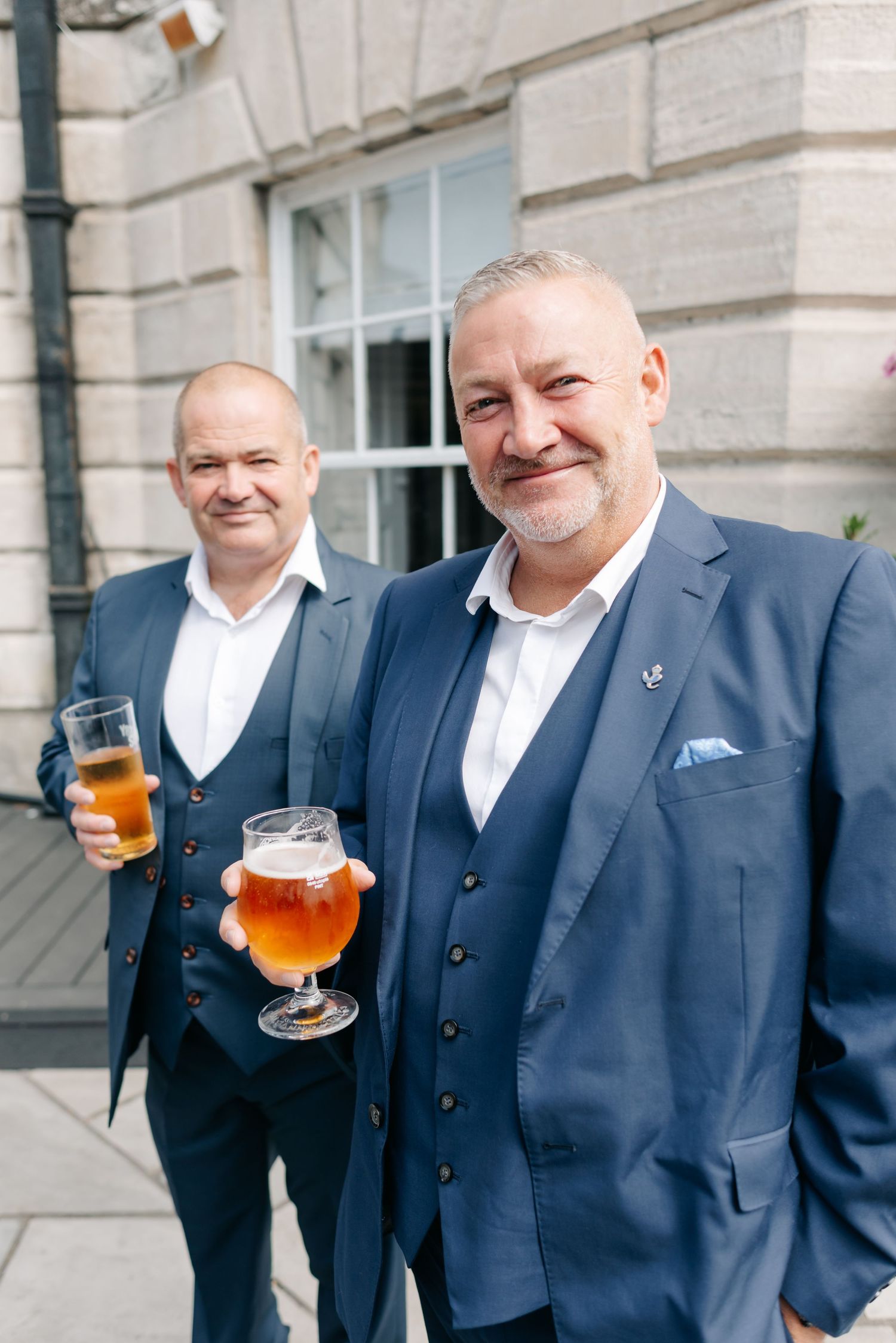Wedding party members in navy suits toast with glasses of beer outside a stone building.