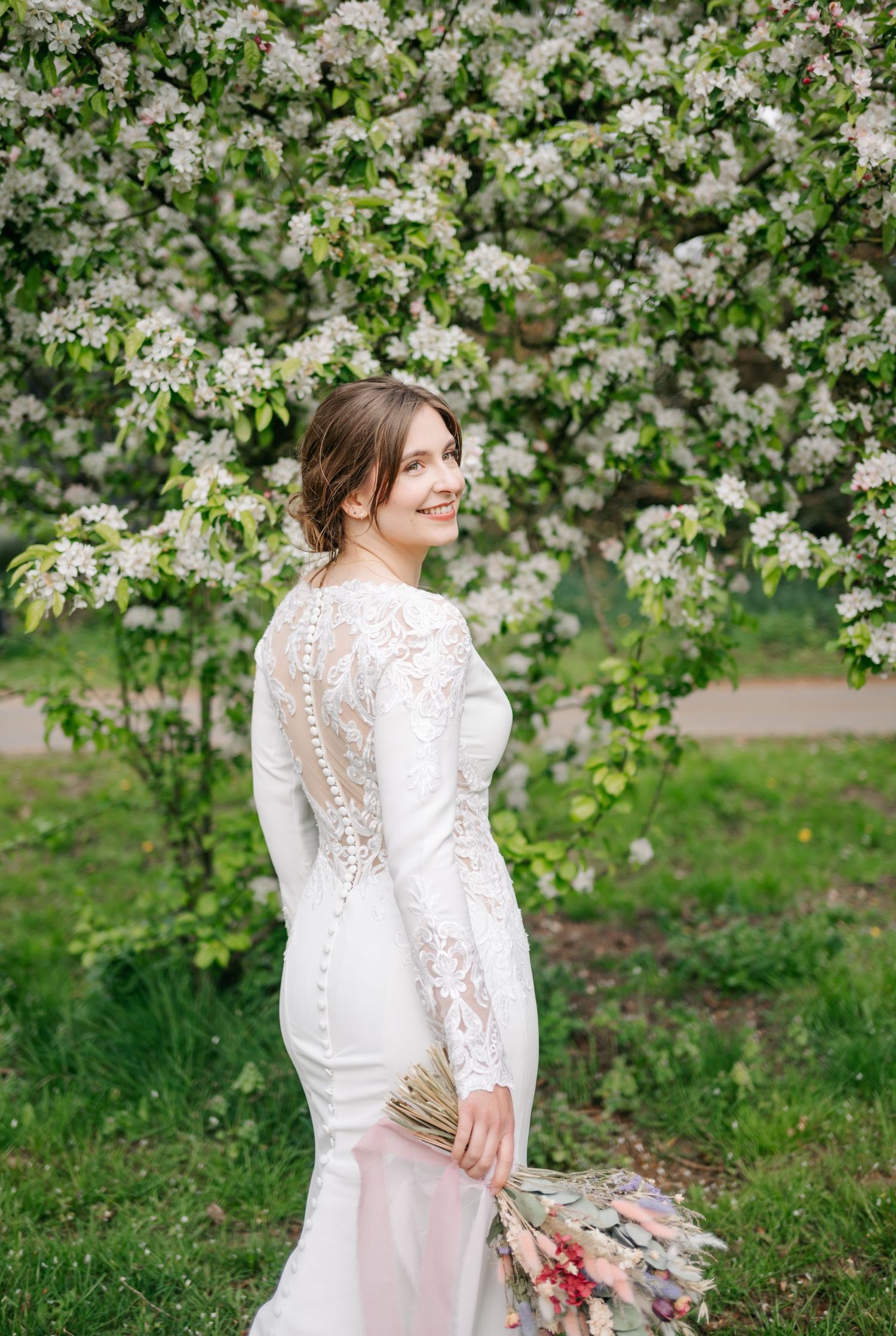 Elegant bridal portrait showcasing lace back detail of wedding dress against blooming spring trees.