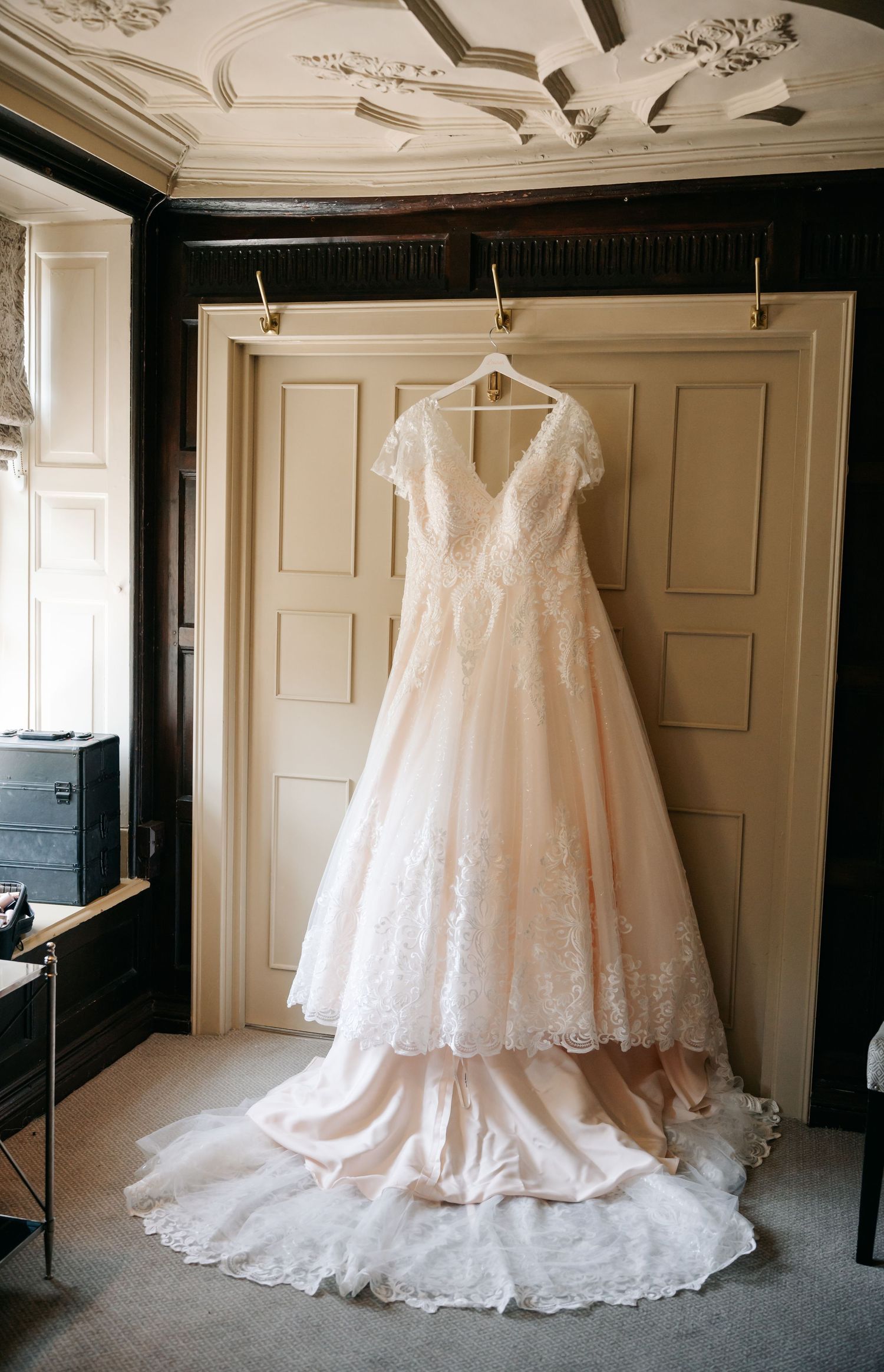 Elegant blush lace A-line wedding dress hanging on a door in a classic ornate room.