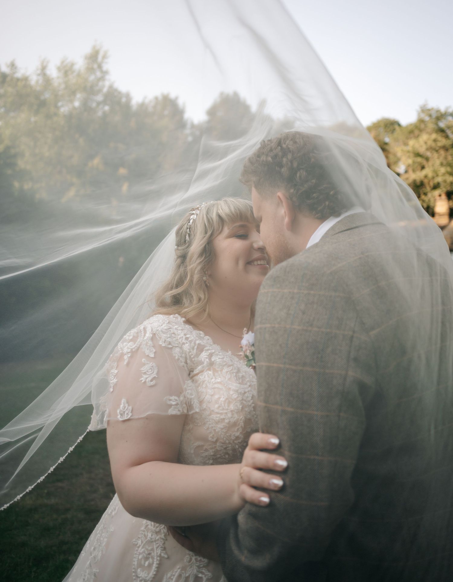 Bride and groom sharing romantic moment under flowing veil in golden hour outdoor wedding photo.
