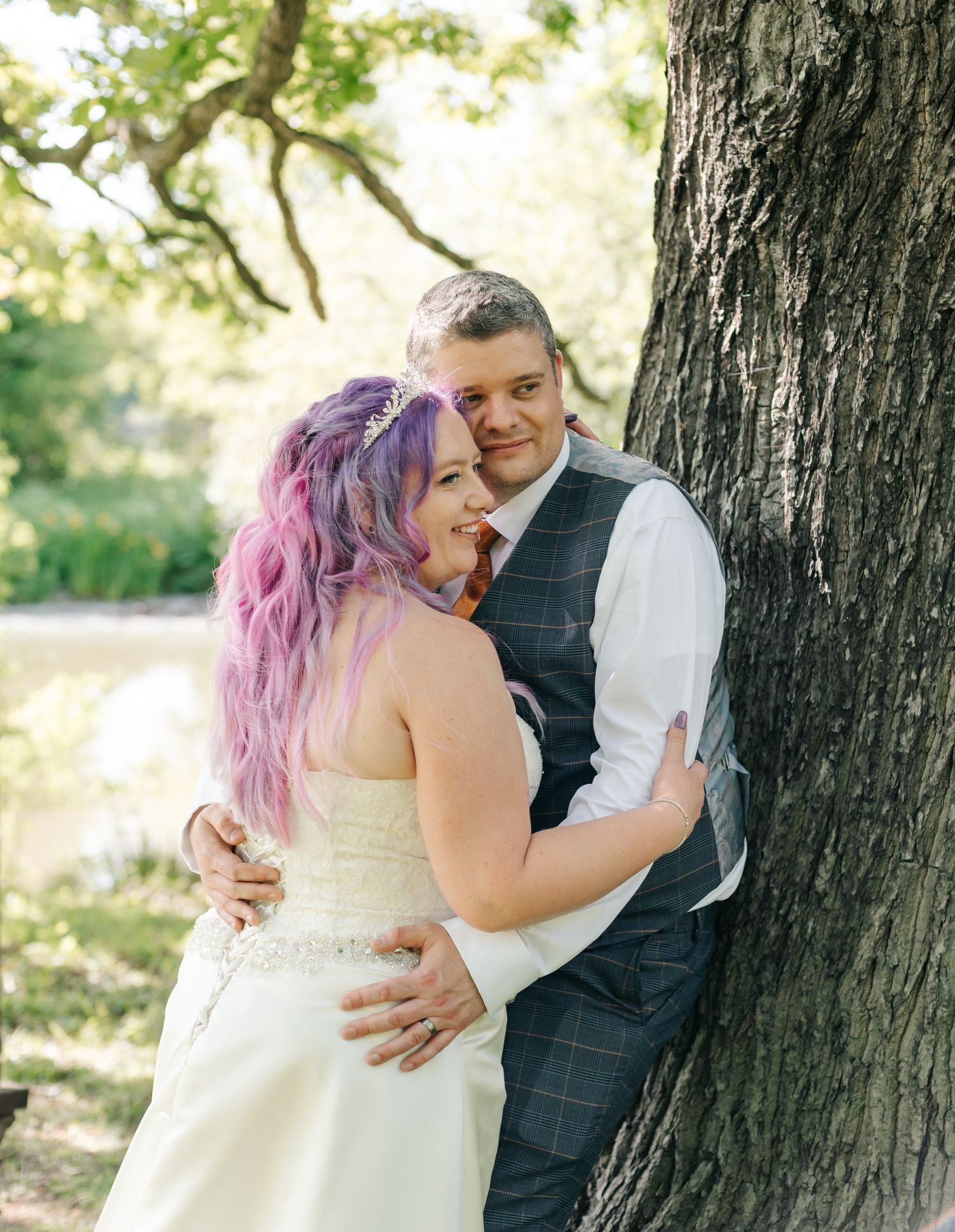 A bride with purple hair and groom in vest embrace by a tree outdoors at their wedding.
