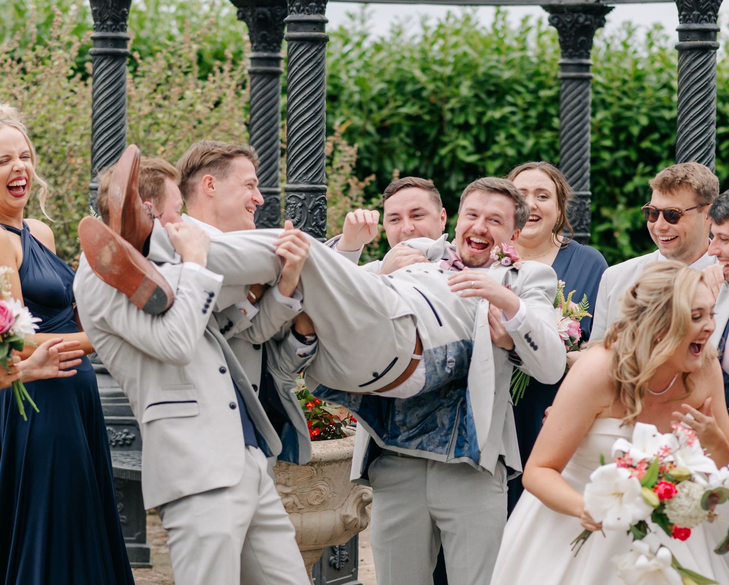 Joyful wedding party celebrating outdoors near decorative black pillars in a garden setting.