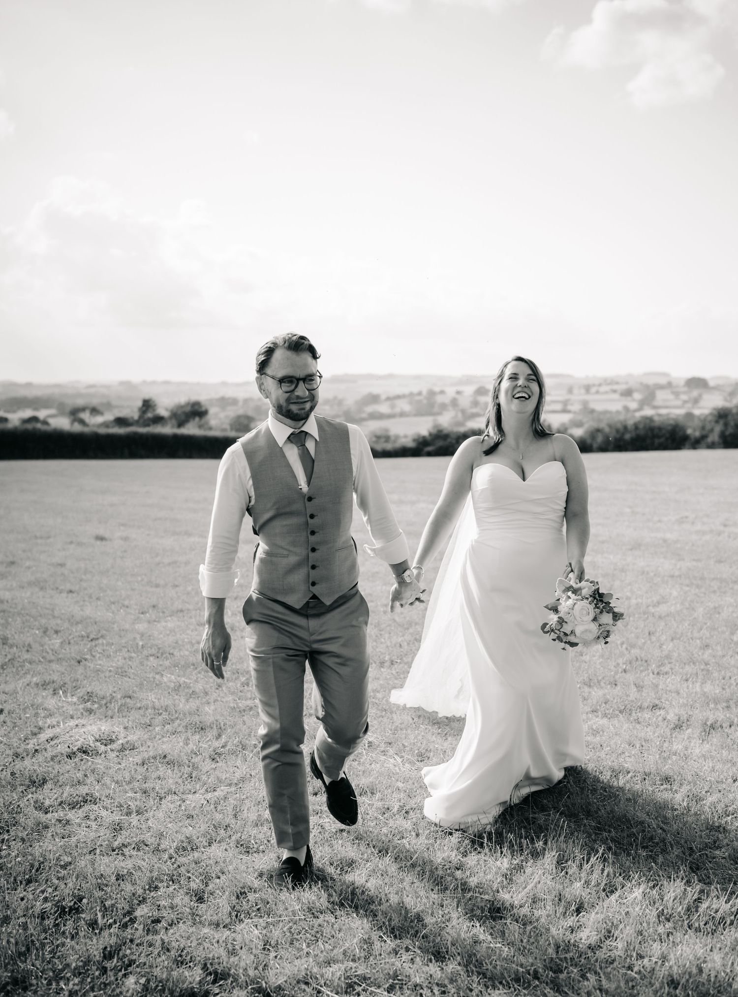 Black and white photo of a laughing bride and groom holding hands walking through a field on their wedding day.