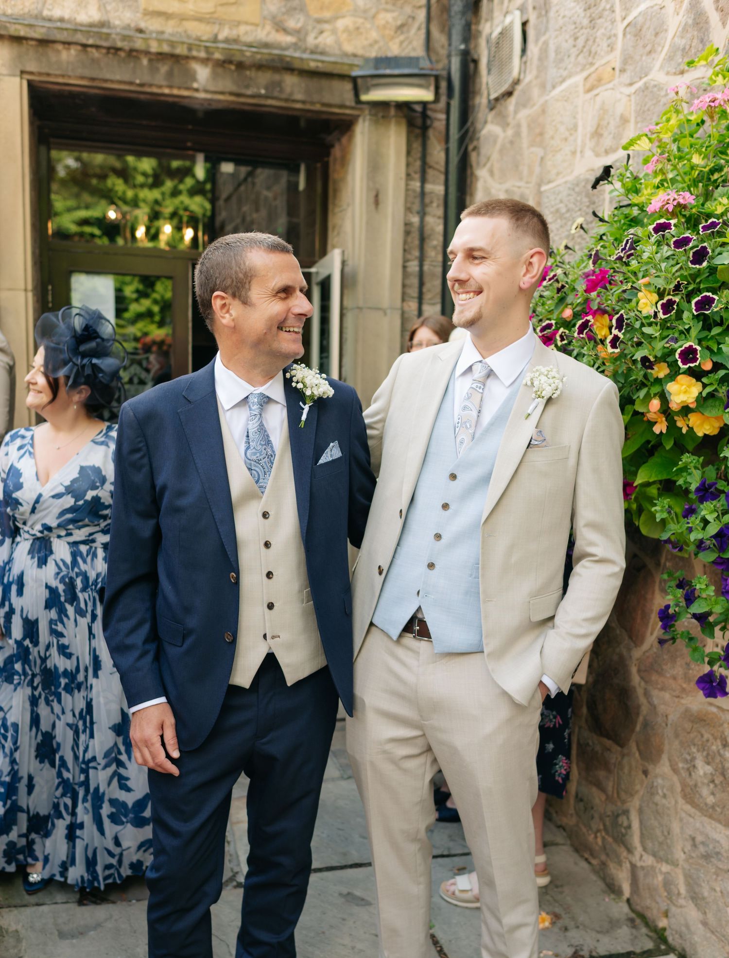 Wedding party members in light beige and navy suits gather outside a stone building with purple flowers.