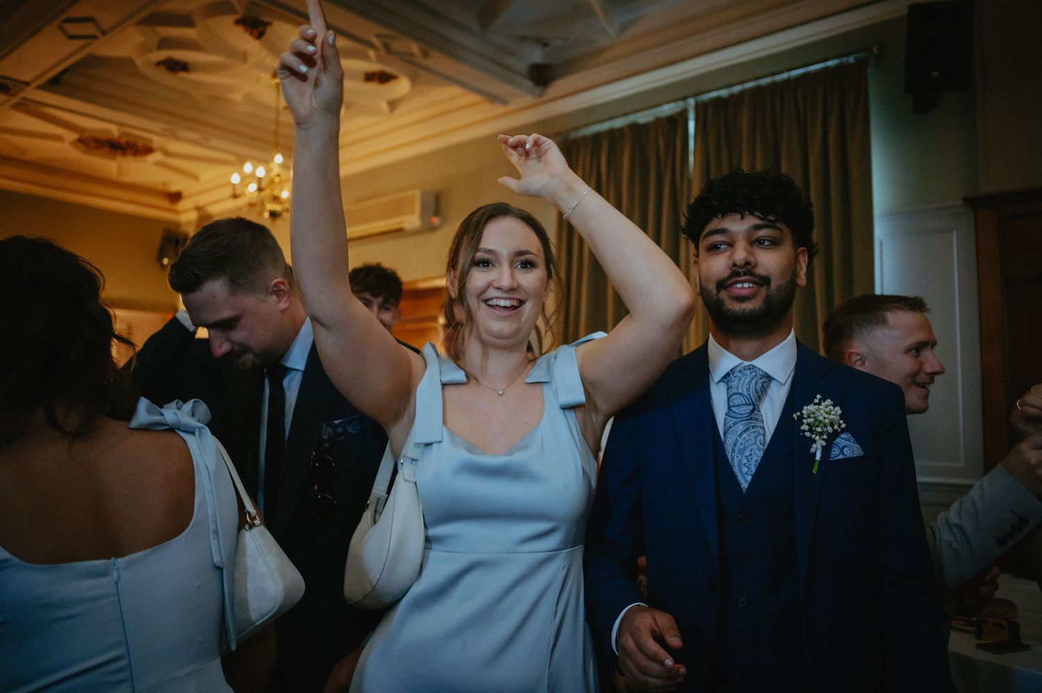Wedding party dancing and celebrating in an elegant ballroom with chandelier lighting.