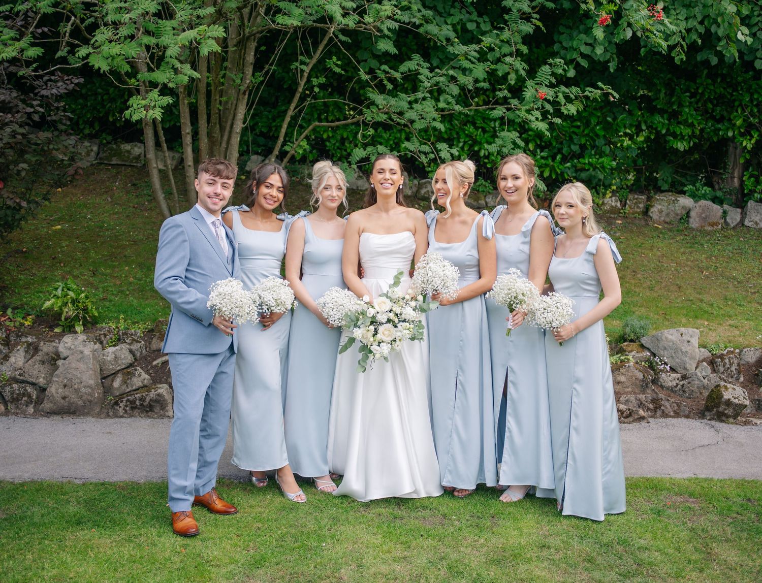 A wedding party wearing light blue dresses and a gray suit poses together in a garden setting.