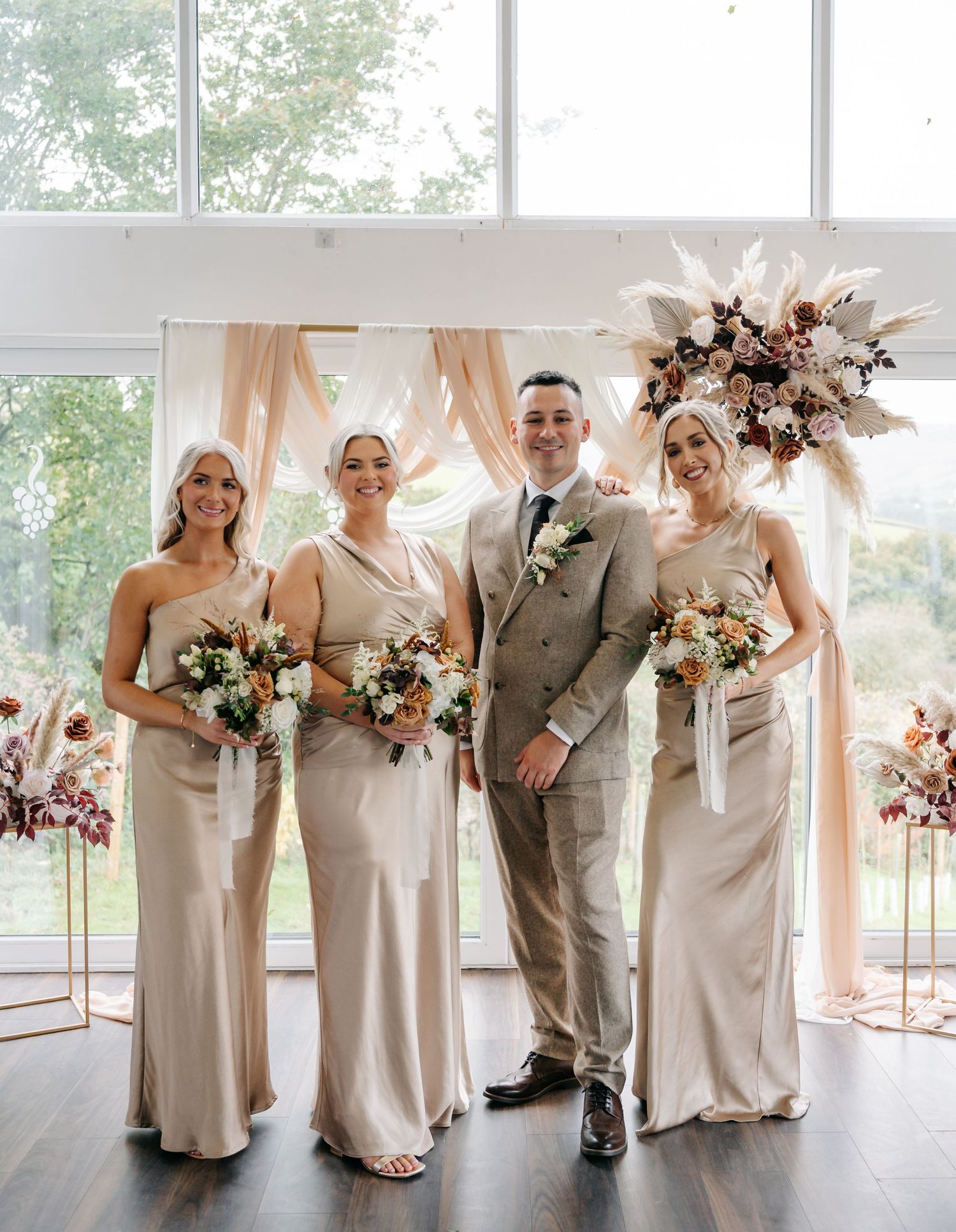 Wedding party in neutral tones posing by large windows in an elegant white venue.