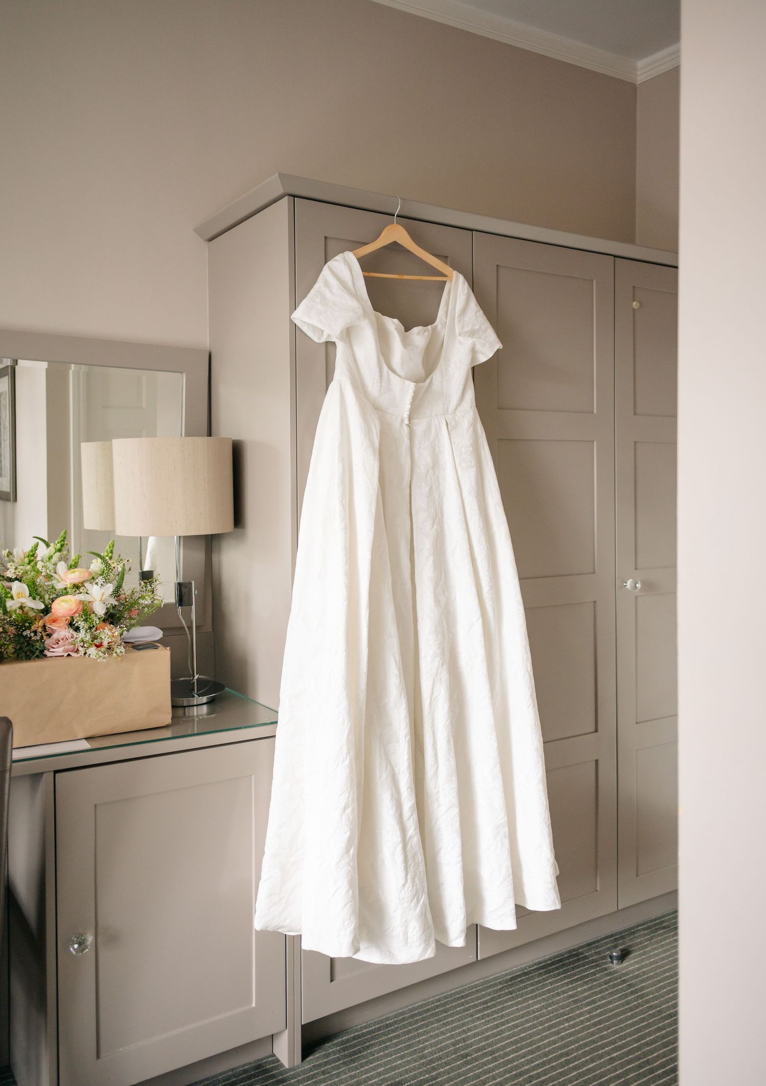 White wedding dress hangs elegantly on a wooden hanger against a neutral grey wall in a hotel room.