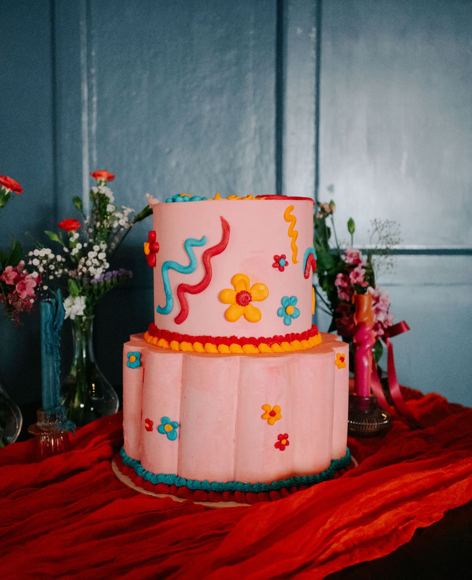 Pink tiered wedding cake with colorful flower decorations on red tablecloth.