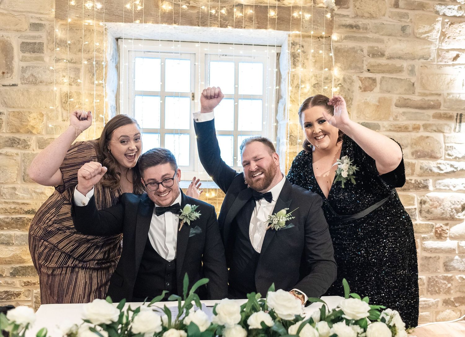 Wedding party celebrates at head table with joyful expressions and raised arms in rustic stone venue setting.