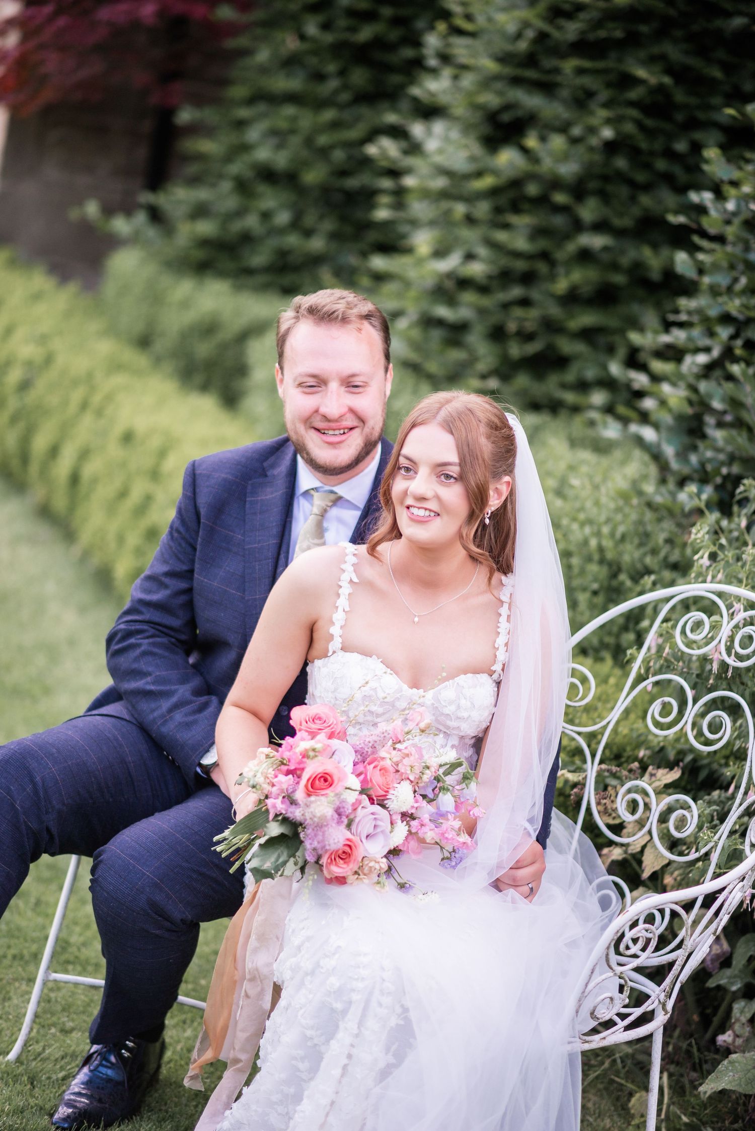 Newlyweds share moment on decorative garden bench surrounded by greenery.