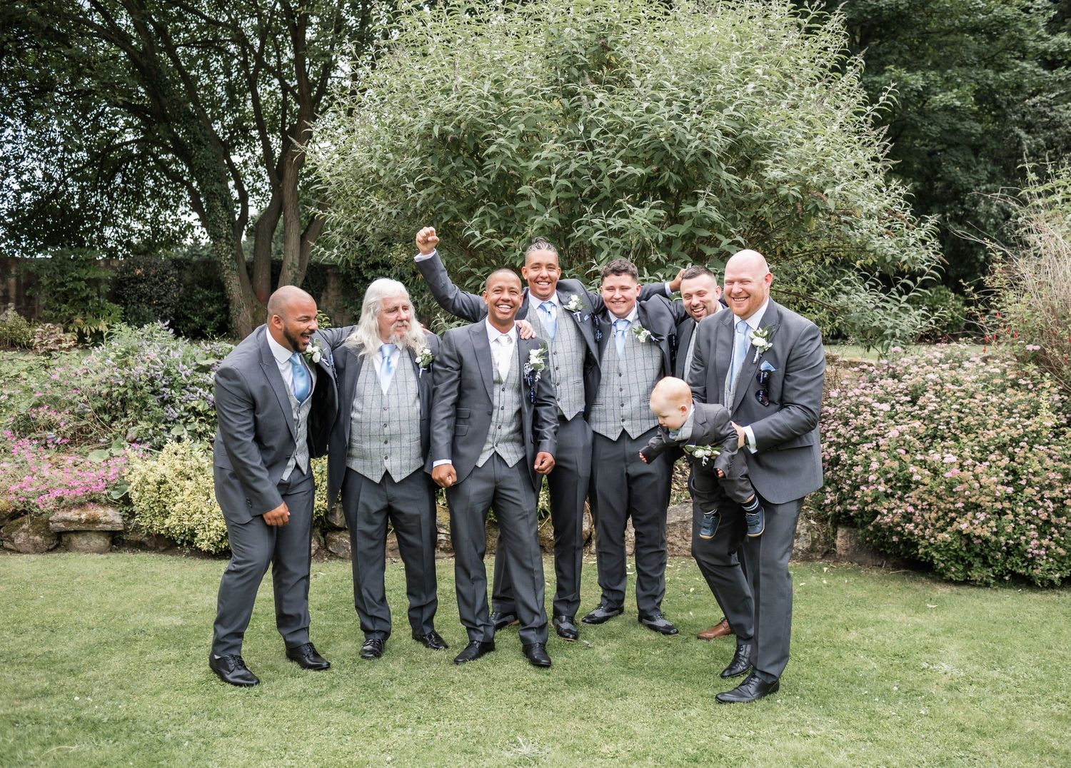 Wedding party in matching grey suits share a candid moment while posing in a garden.