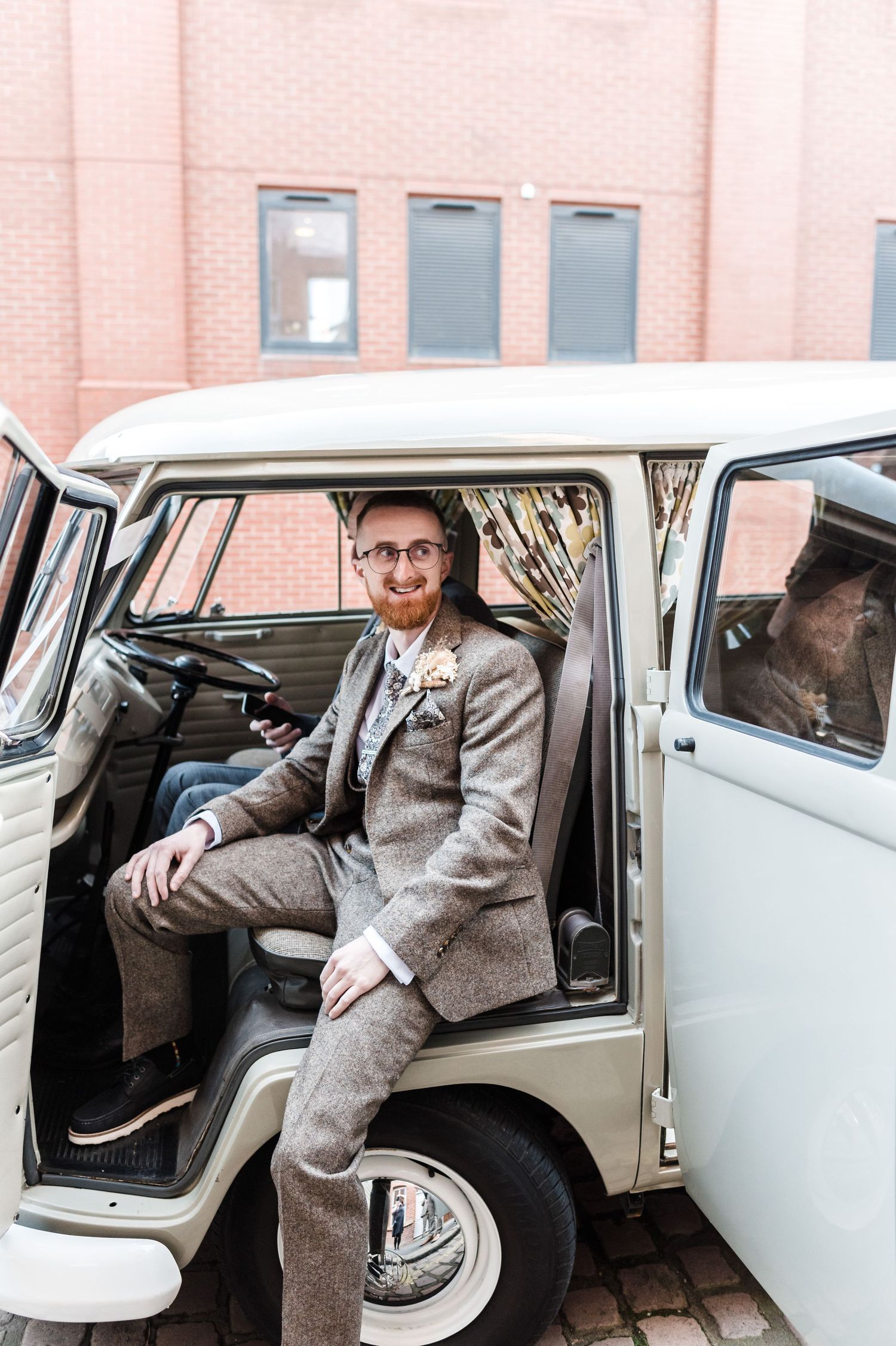 A person in a gray tweed suit sits in a classic white vintage van with the door open.
