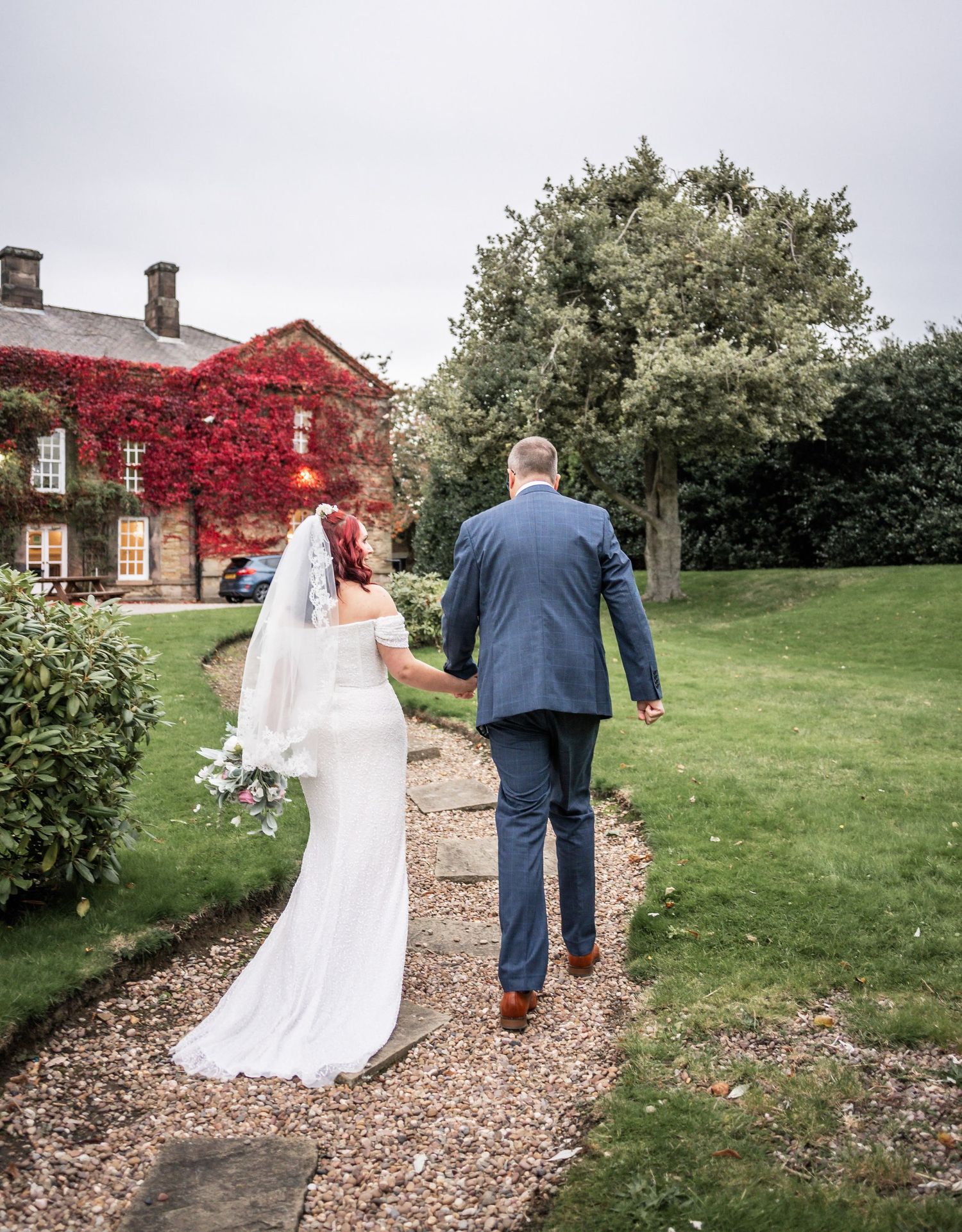A couple walks down a garden path towards a historic building with red ivy-covered walls.