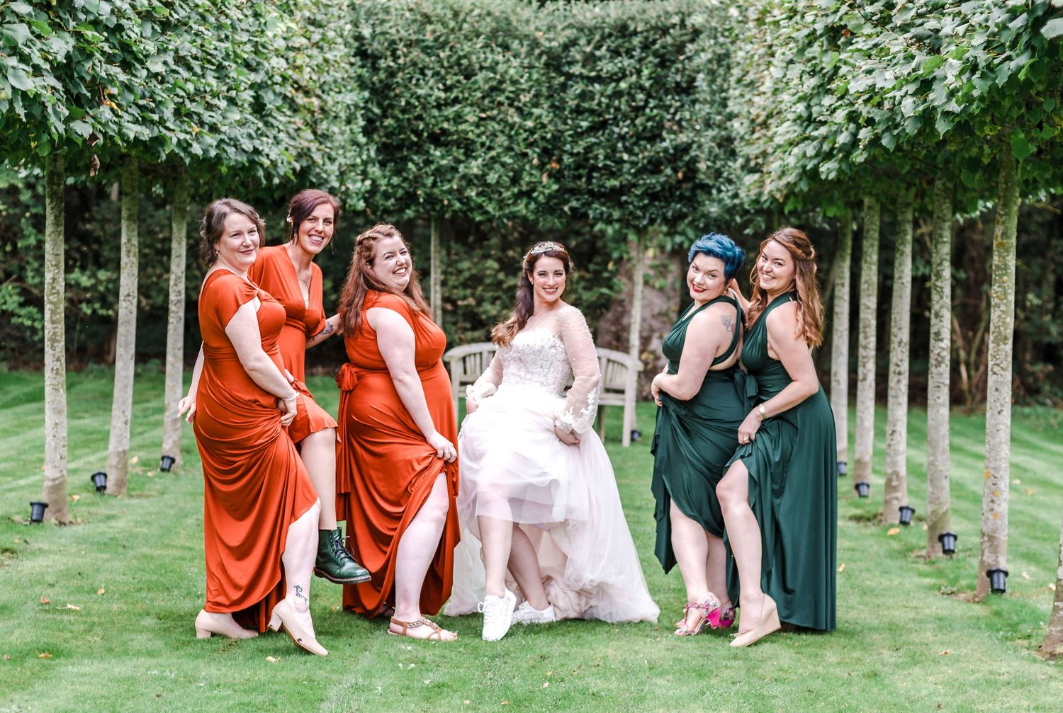 Bridal party in orange and green dresses pose playfully between hedged garden path.