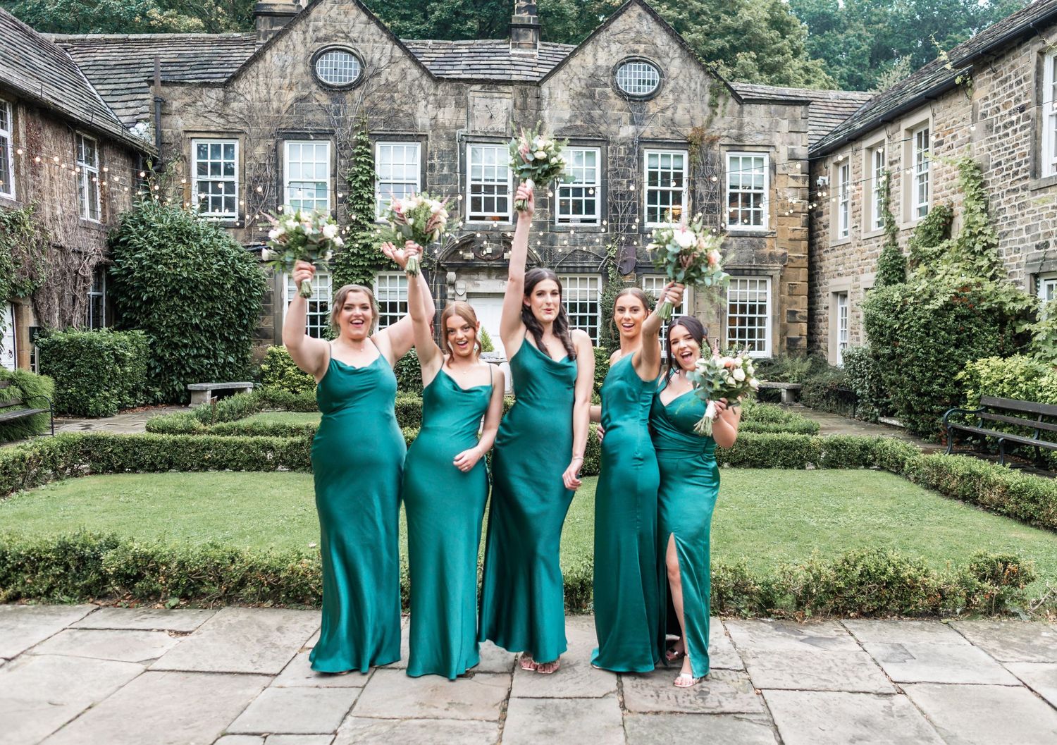 Bridesmaids in matching emerald green dresses celebrating in front of stone manor house.